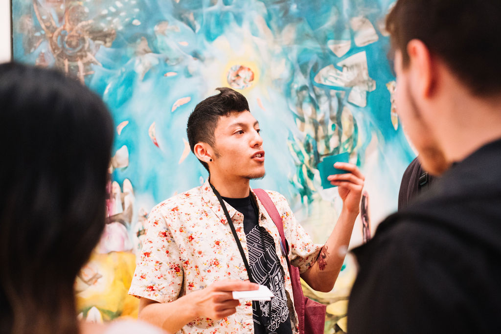 A tour guide with black hair speaks in front of landscape painting with a deep blue sky, a bright multicolored sun with a yellow aura, and abstract figures.