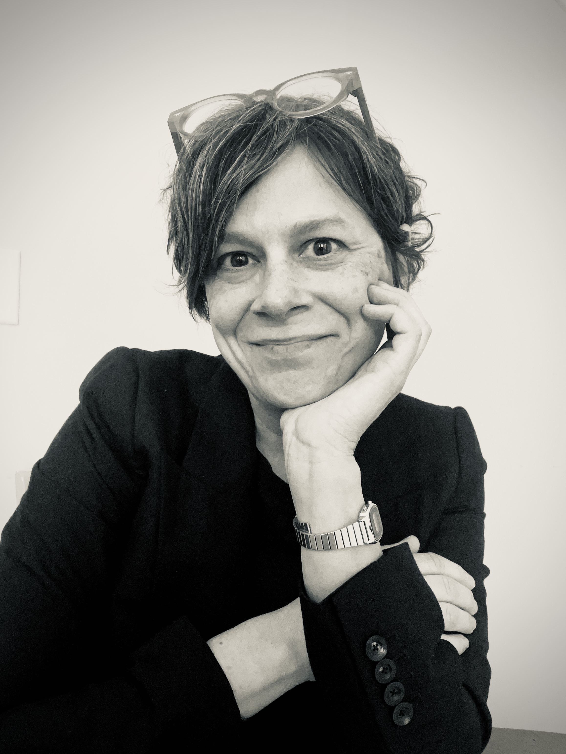 Smiling woman with short hair, wearing glasses on head and a dark blazer. She rests her chin on her hand, exuding a friendly and relaxed demeanor.