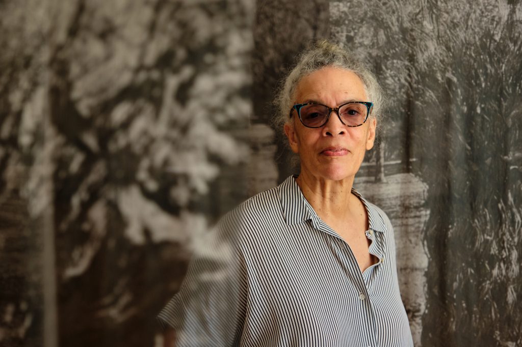 A light skinned black woman with close-cropped gray hair, black framed glasses, and a gray shirt stands before transparent gray and white tapestries evoking foliage.