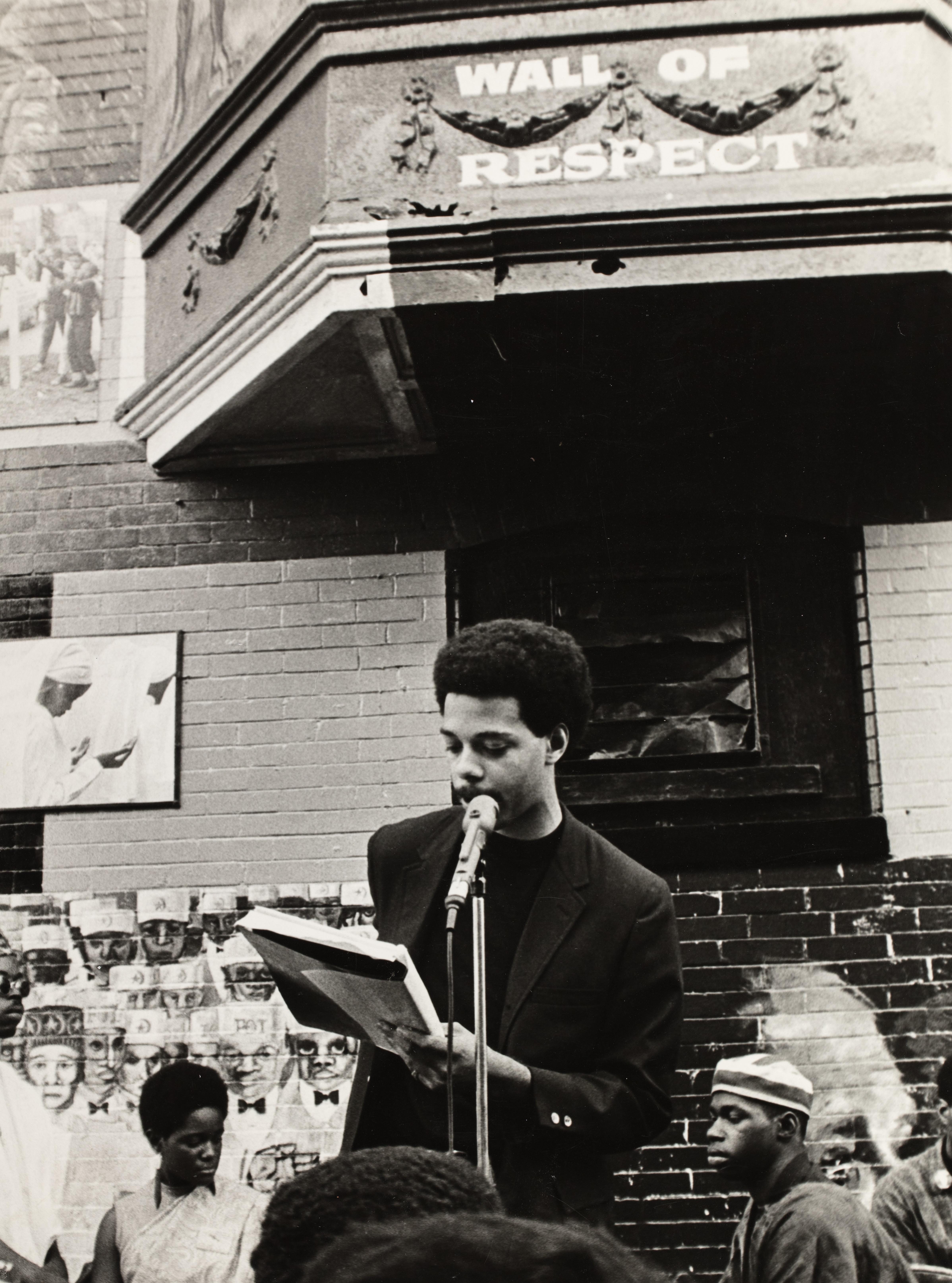 Black-and-white photograph of a man reading from a notebook into a microphone while standing in front of a brick building. He wears a dark jacket and is positioned beneath a sign that reads “WALL OF RESPECT.” Behind him are large painted portraits and photographic collages featuring Black cultural figures. Two seated individuals are partially visible near the wall, listening intently. The crowd in the foreground frames the speaker, emphasizing a communal setting and outdoor public gathering.