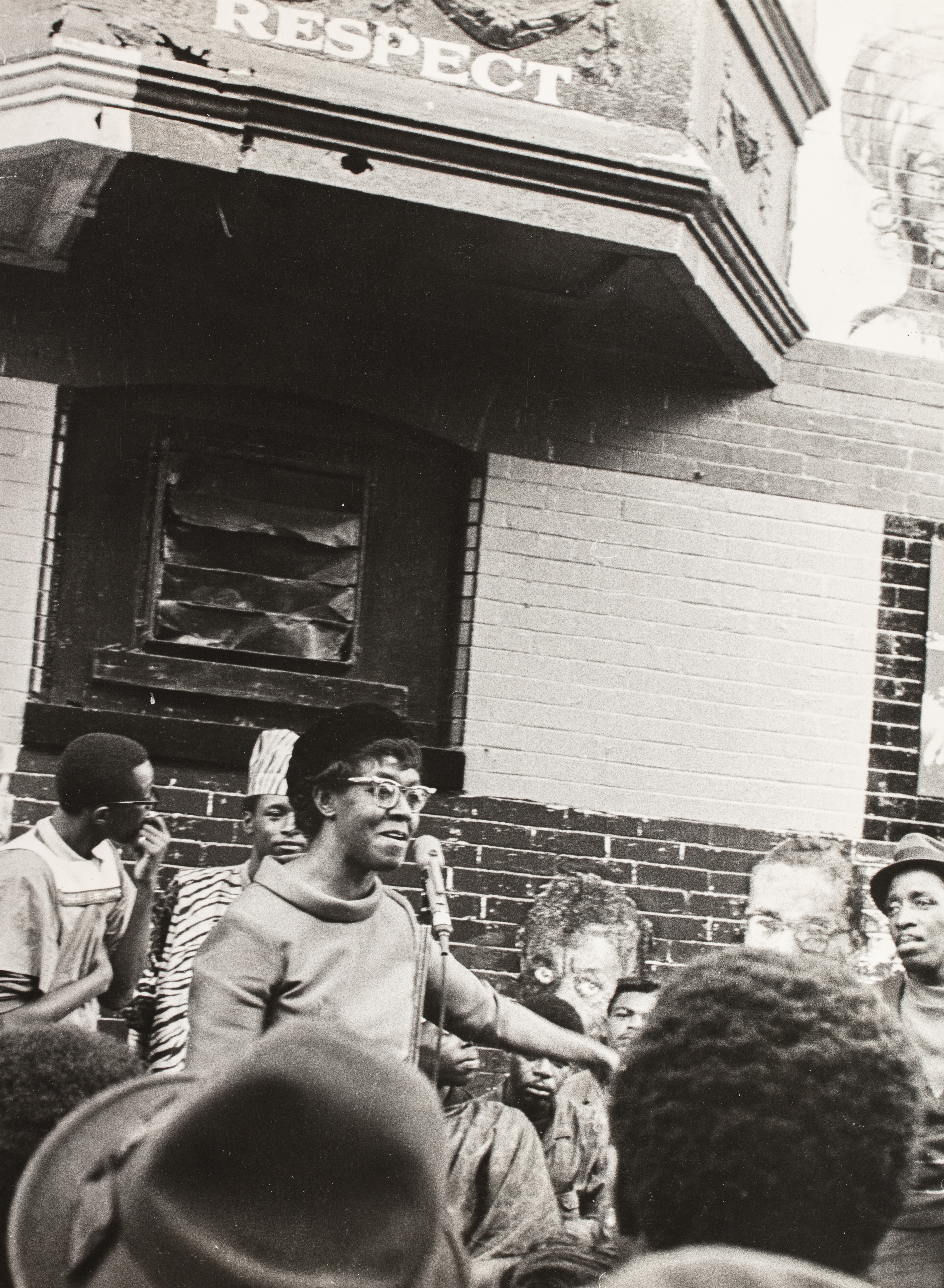 Black-and-white photograph of a woman speaking into a microphone in front of a mural-covered brick building. She wears a hat and glasses and gestures with one arm extended. A group of people, mostly men, stand and sit around her, some in patterned clothing and traditional hats. Above her head, part of a mural reads “RESPECT.” Painted portraits and partial faces appear behind the group, integrated into the wall. The crowd in the foreground is closely gathered, suggesting an engaged public audience.