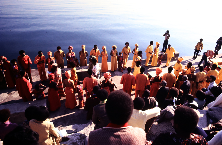 Color photograph showing a large group gathered at a lakeside for what appears to be a ceremonial event or performance. The scene is viewed from behind a seated audience on concrete steps, facing a wide semicircle of participants dressed in coordinated outfits. Most wear garments in shades of yellow, orange, and red, including robes, dresses, and head wraps. A few individuals hold cameras, including a person at the far right capturing the event. The calm water of the lake fills the background, emphasizing the outdoor setting and serene atmosphere.