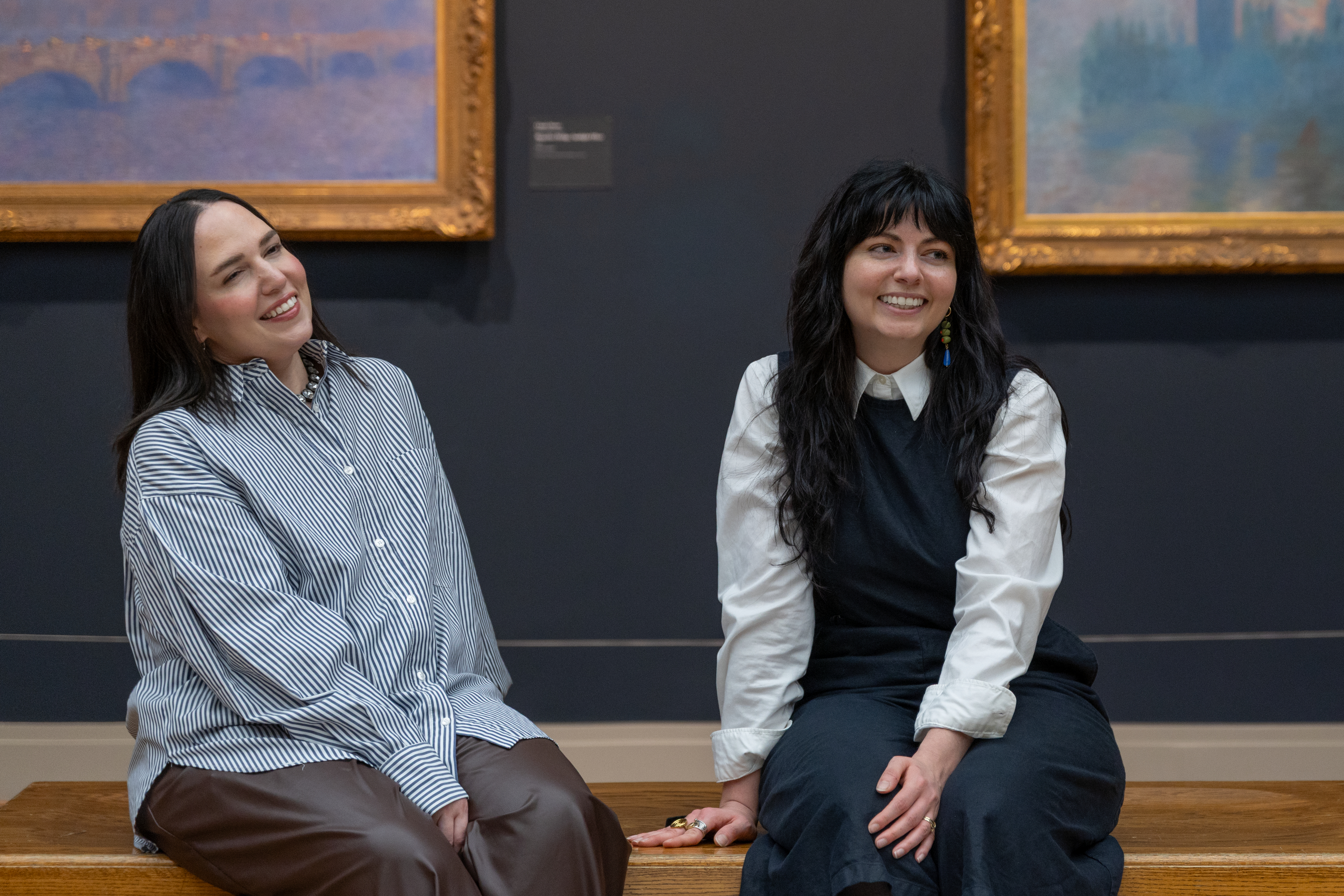 A color photograph shows two women with long dark hair, Zahra Bahia and Sam Grassi, seated on a wooden bench at center against gold-framed blue-toned paintings by Monet that hang on a dark wall behind them.