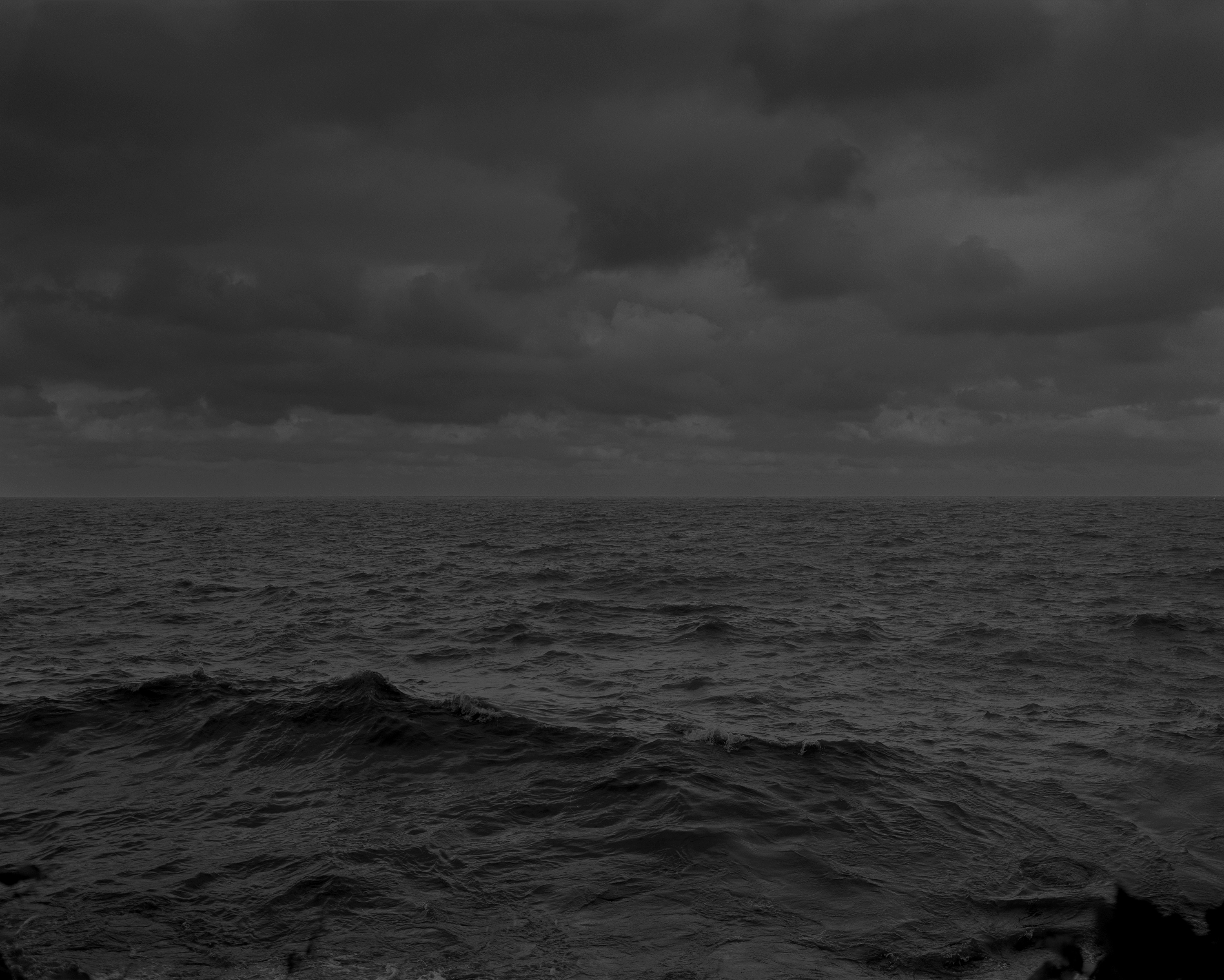 A dimly lit, dark black-and-white photograph of a moderately rough sea with low waves and a sky densely packed with clouds. The sea and sky each take up half of the image. 