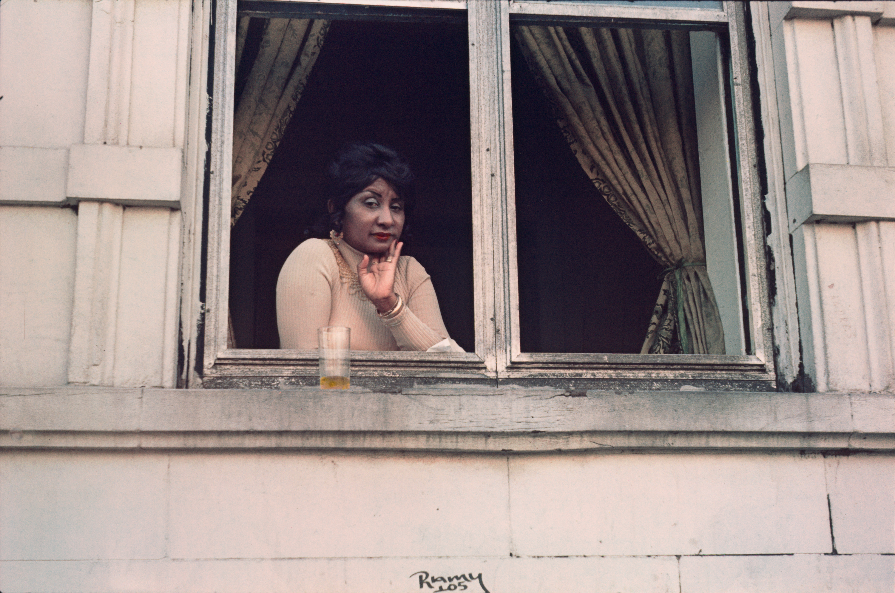 A medium-skinned woman with dark hair, red lipstick, jewelry, and a beige turtleneck looks out a big window with a cup on the sill, eyebrow raised, skeptical.