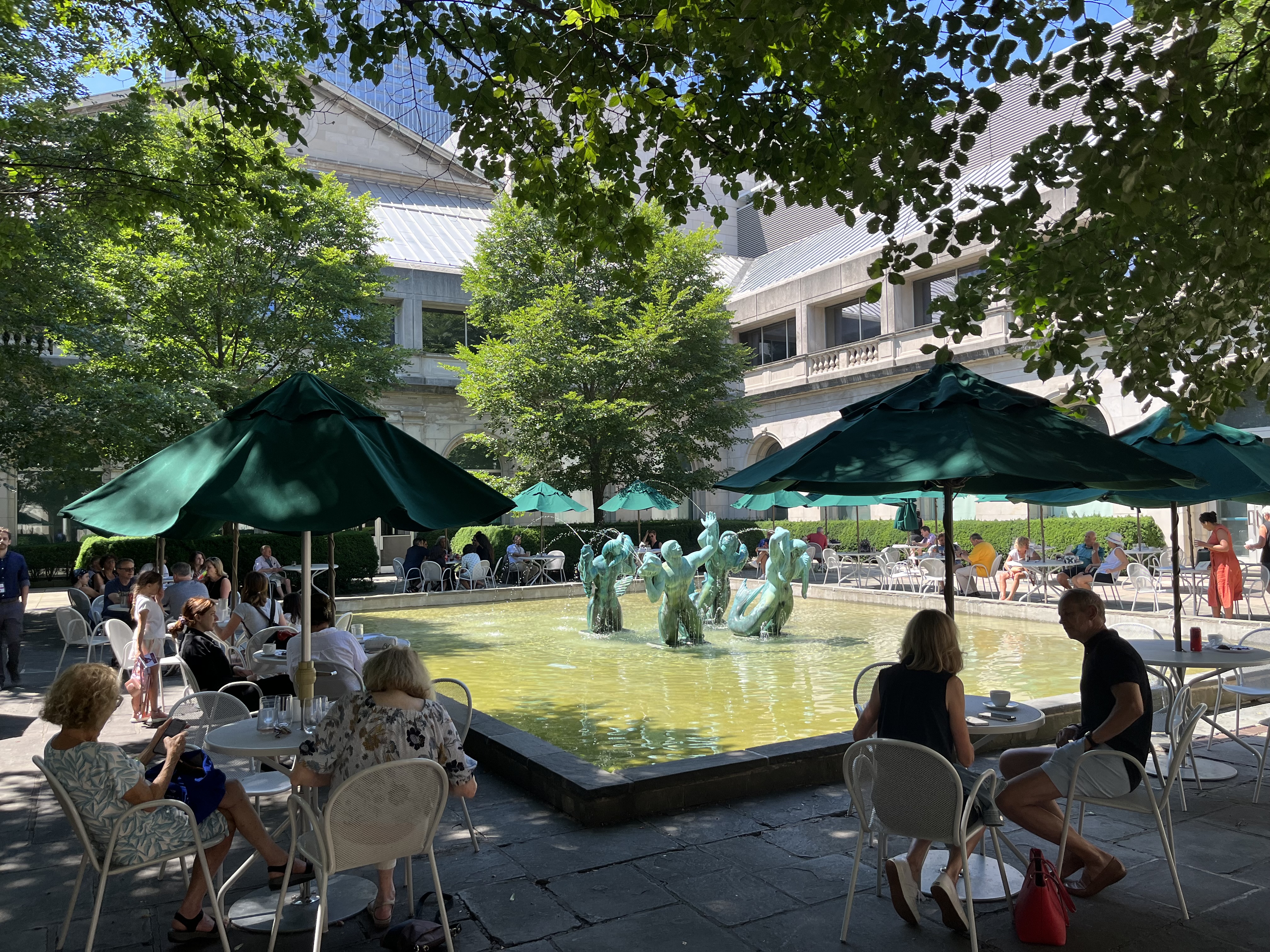 Photo shows an outdoor space—a courtyard—with a central shallow pool and fountain of four figures with green patina. Surrounding the pool are many bistro tables with large green umbrellas. People dine in the shade of trees and the surrounding building.