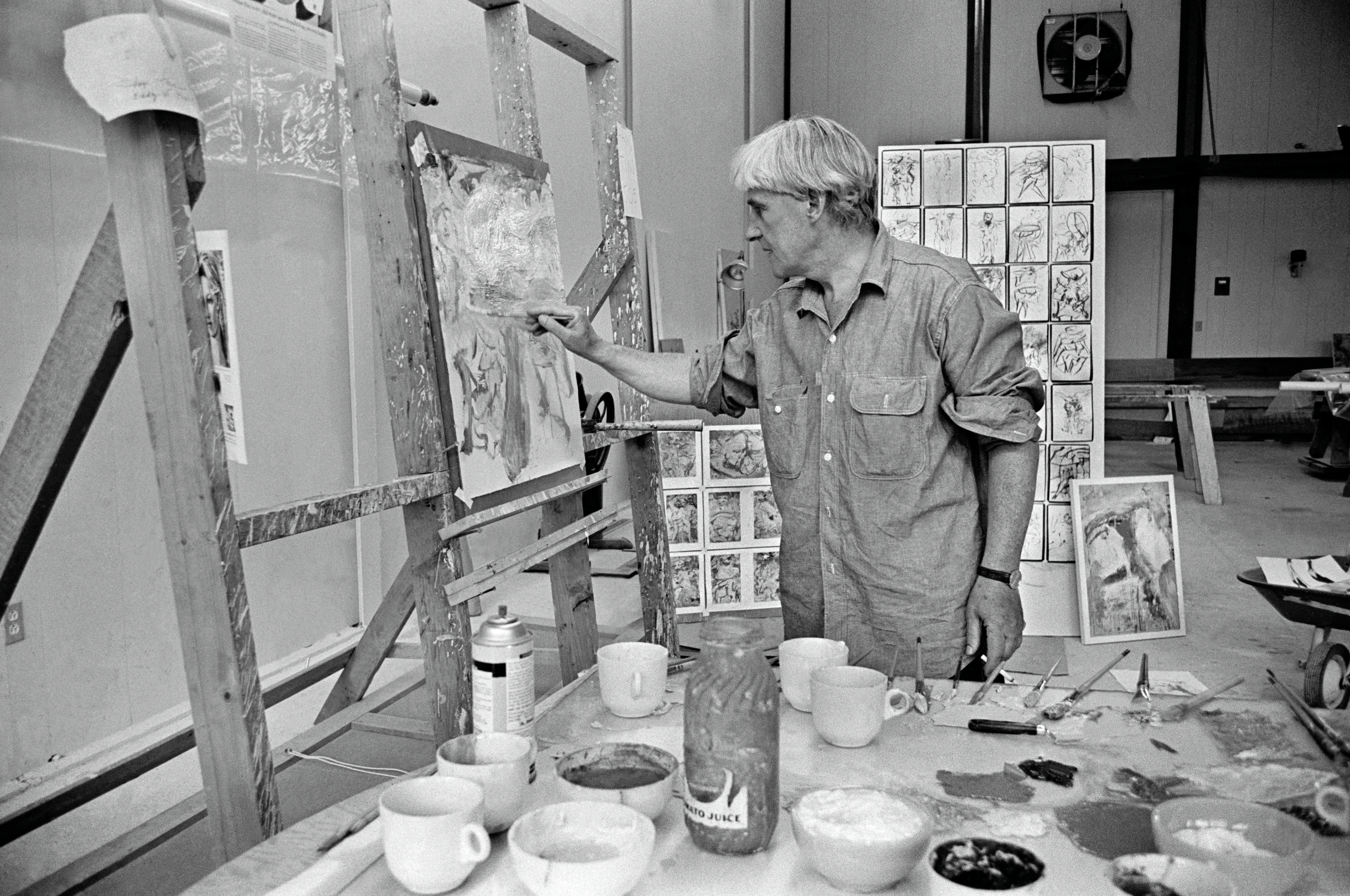 A black-and-white photograph of an art studio shows an adult with short hair at center-right reaching to a canvas on an easel at left, with a foreground table holding cups, jars, brushes, and paint, and a tall grid of sketch pages standing behind at center.