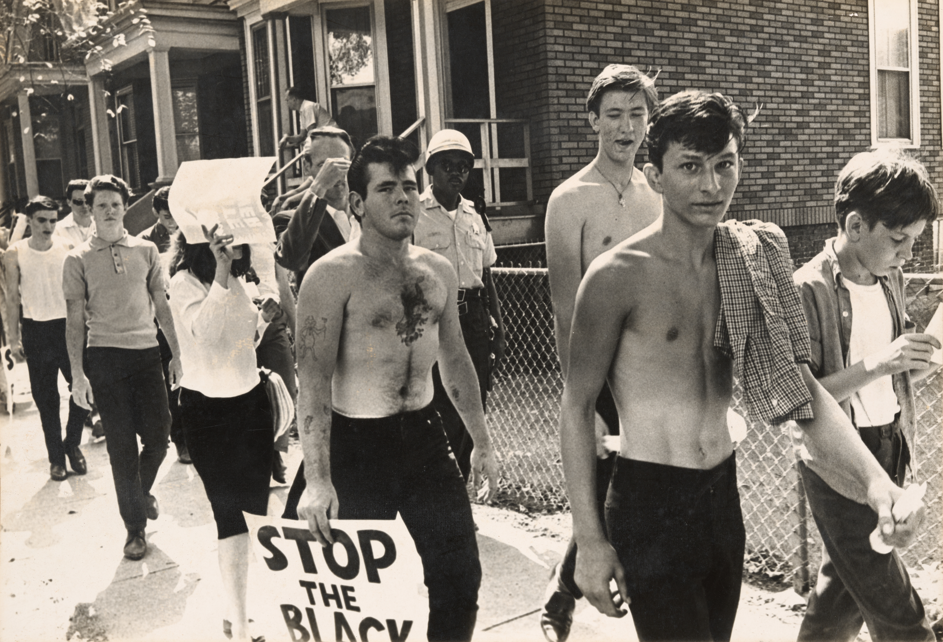 Black-and-white photograph showing a group of young white protesters marching along a sidewalk in a residential neighborhood. Several shirtless young men lead the group, one holding a sign that reads "STOP THE BLACK" with additional text obscured. Behind them, more marchers follow, including a woman shielding her face with a newspaper. A uniformed police officer stands in the background near a brick building, watching the procession. The expressions of the marchers vary, some looking serious, others indifferent or defiant. The mood suggests racial tension and protest.