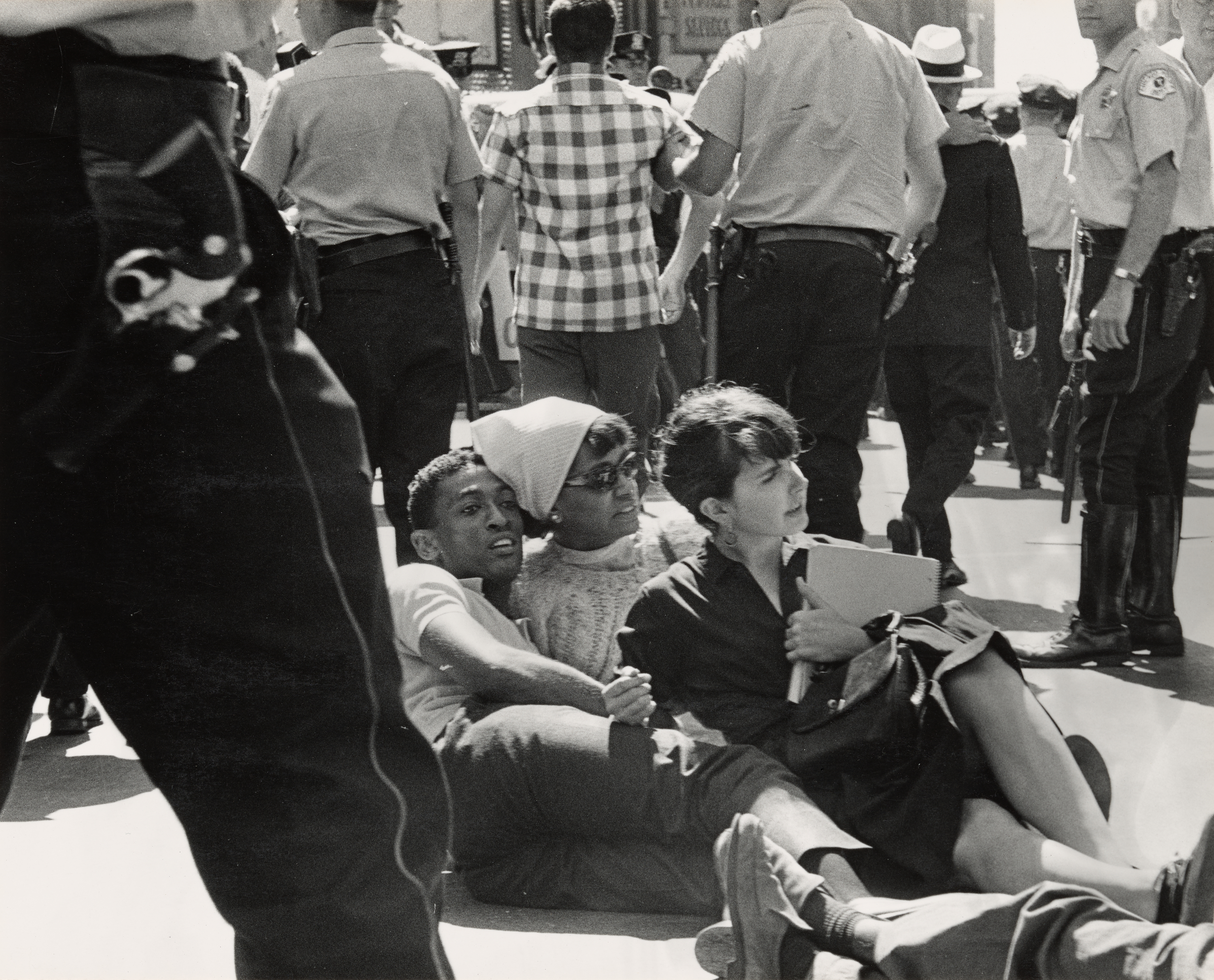 Black-and-white photograph showing a group of young civil rights protesters sitting on the ground in the middle of a street. Three people are clearly visible in the foreground, sitting close together: a young Black man, a woman in a light sweater and headscarf, and a woman in a dark dress holding a notebook. All appear calm and composed. Around them, uniformed police officers walk and stand in formation, with their backs turned or partially visible, creating a sense of tension. The scene conveys a peaceful protest surrounded by law enforcement presence.