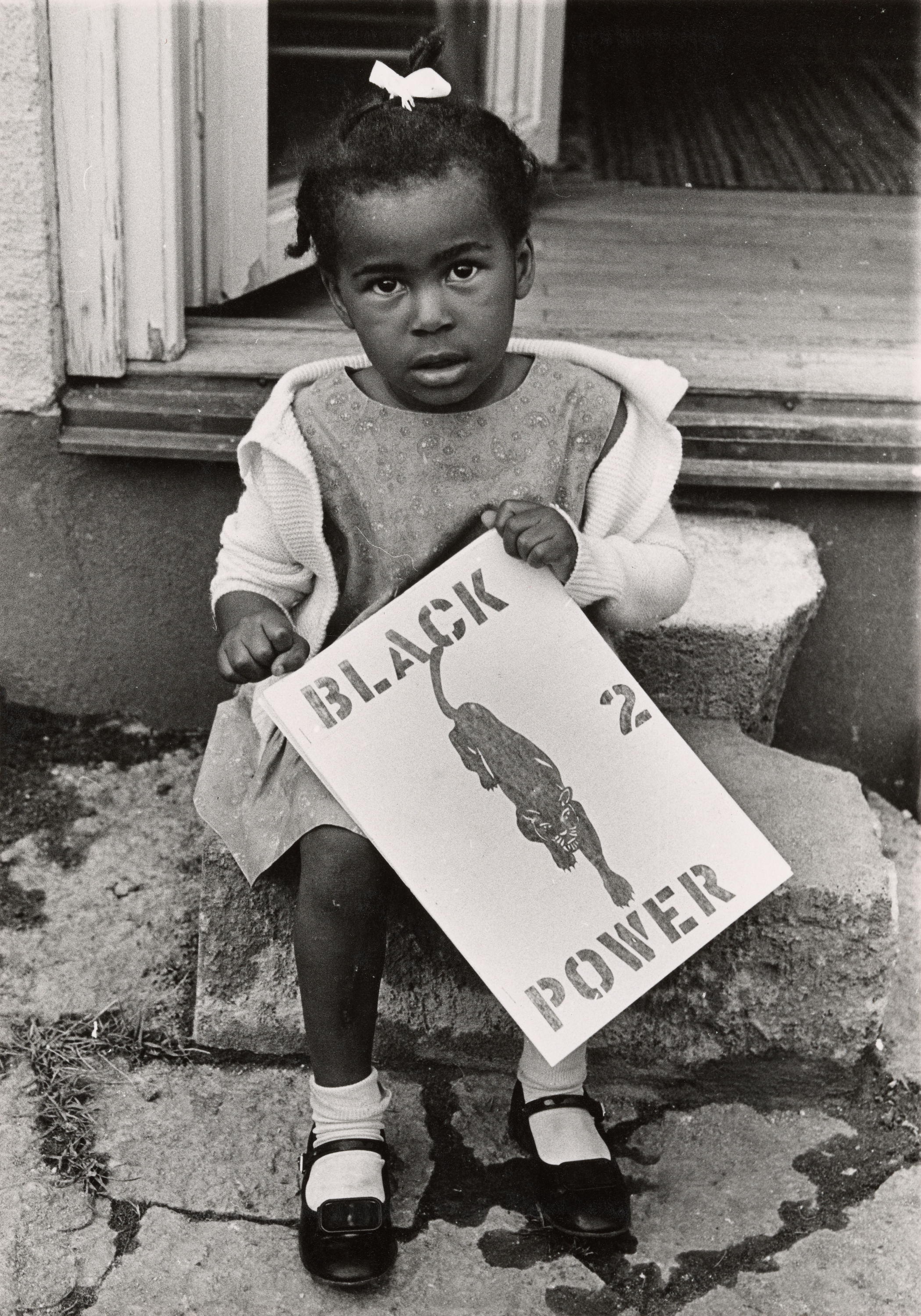 Black-and-white photograph of a young Black girl sitting on concrete steps in front of an open doorway. She wears a light cardigan over a patterned dress, white socks, and black Mary Jane shoes. A bow is tied in her hair. She holds a large sign in her lap that reads “BLACK POWER” with a stenciled image of a panther. The number “2” is placed between the words. The girl's wide-eyed expression meets the camera directly.