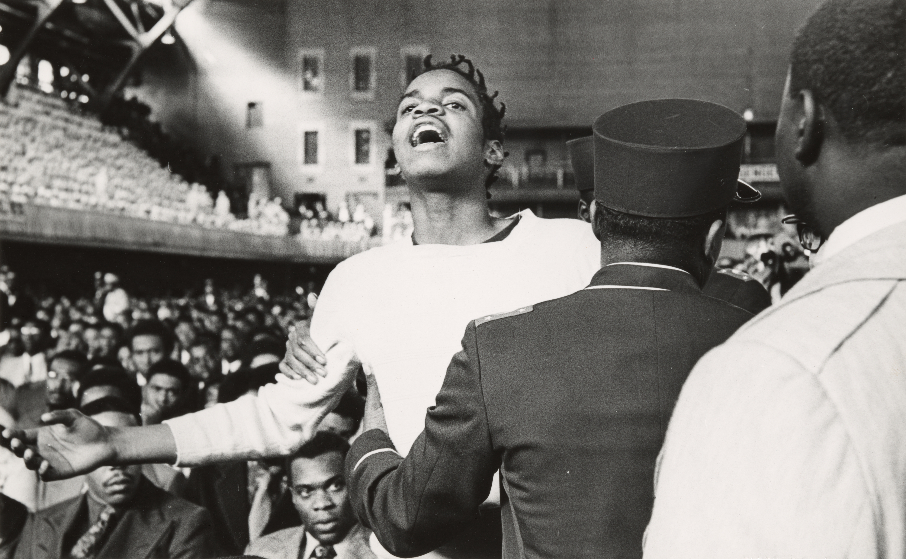 Black-and-white photograph showing a crowded indoor gathering with a person in the foreground raising their arms and tilting their head back, mouth open in what appears to be an emotional outburst or chant. The person wears a light-colored top and has short, twisted hair. Several men in uniform jackets and pillbox-style hats face the person, gently holding their arms. Rows of seated people fill the background, along with a balcony level also crowded with people. The scene takes place in a large auditorium or arena with overhead lighting and visible architectural beams.