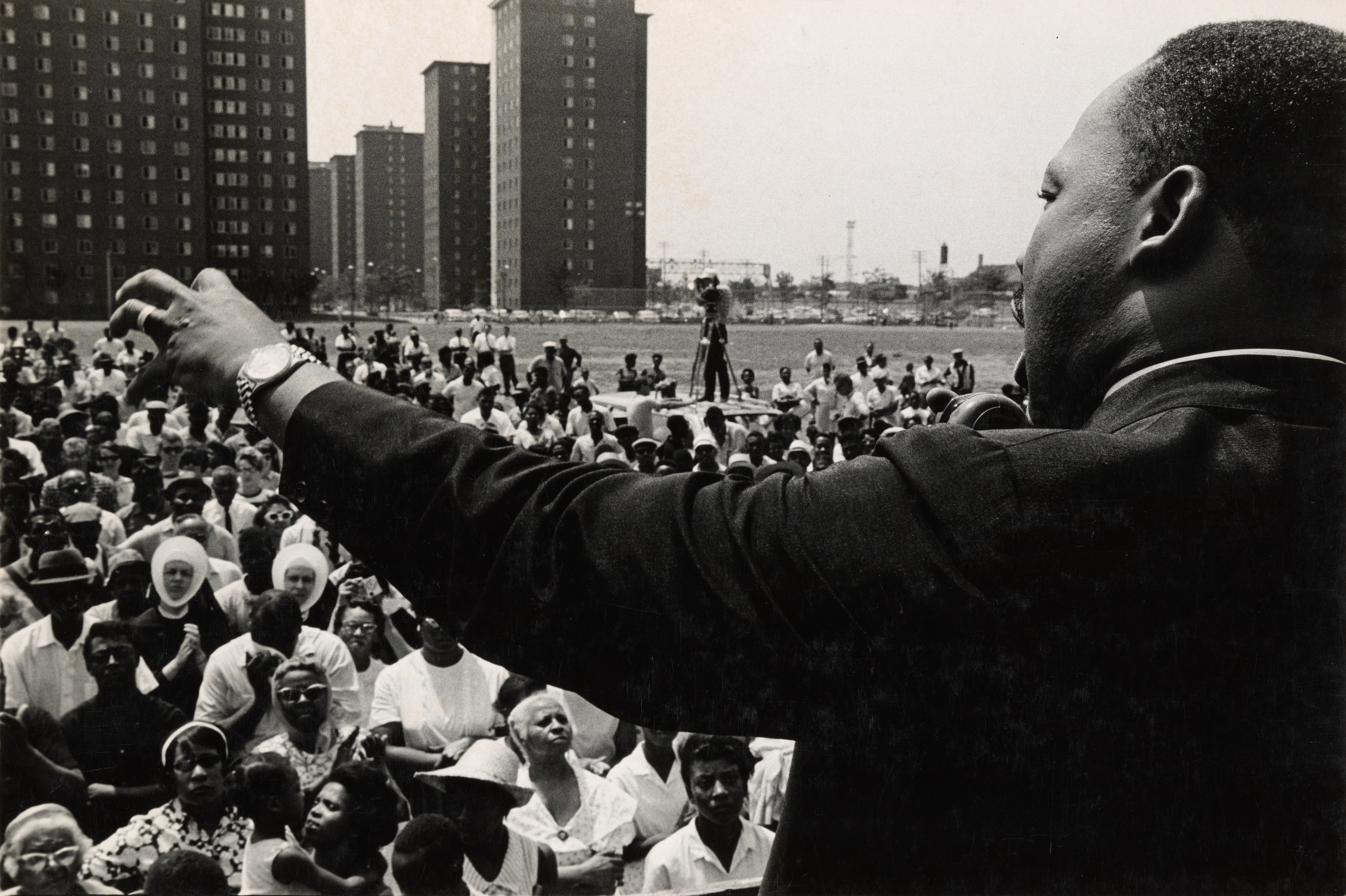 Black-and-white photograph taken from behind and slightly to the side of a man speaking to a large outdoor crowd. His right arm is extended in a gesture, and he wears a dark suit and a watch on his left wrist. A microphone is visible in front of him. The crowd, made up of men, women, and children, faces him, many looking up attentively. Two nuns in white habits are seated near the front. In the background are several tall apartment buildings and an open grassy area. A camera operator stands on a raised platform in the distance.