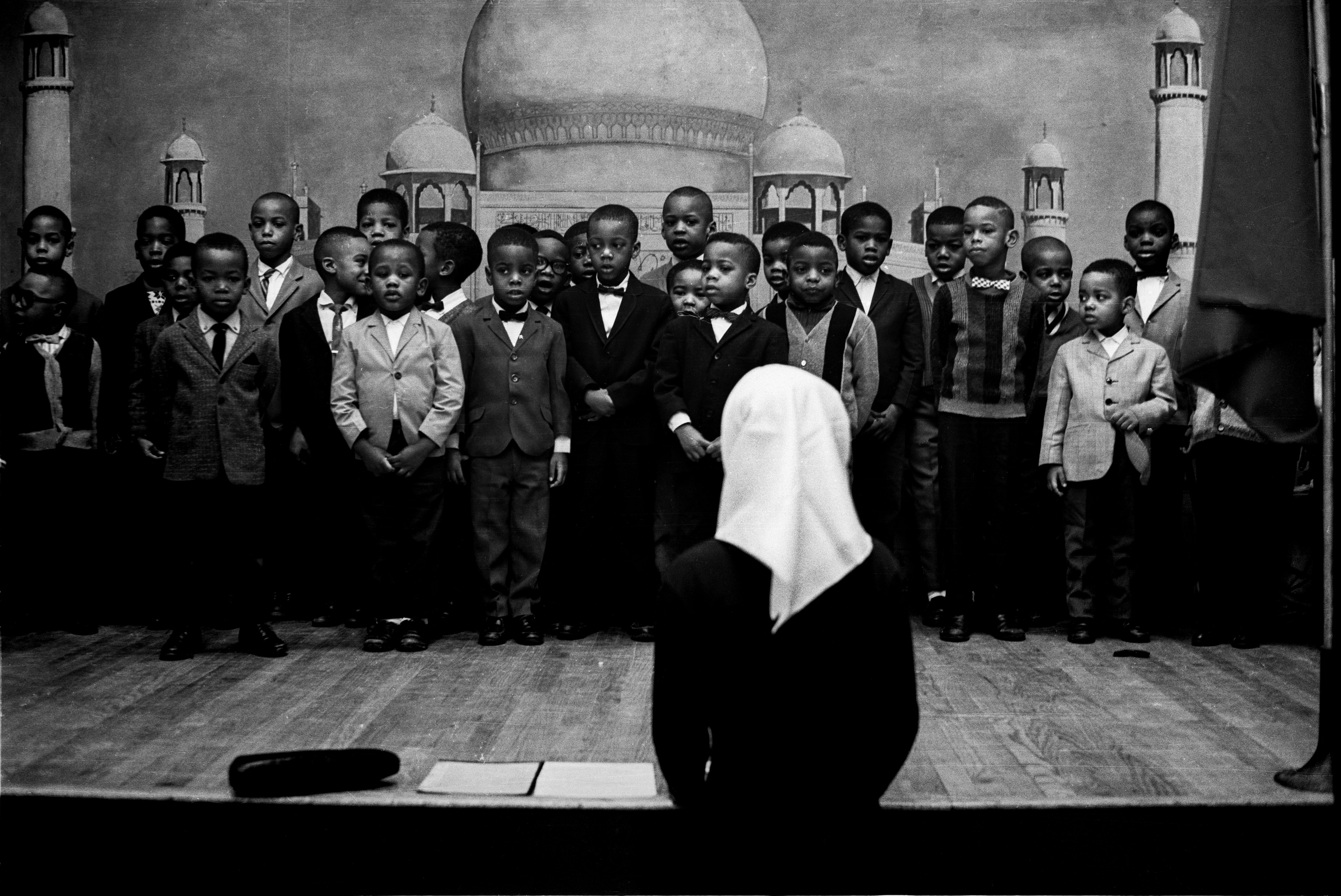 Black-and-white photograph of a group of young boys standing on a stage in several rows, facing forward. The boys are dressed in formal and semi-formal clothes, including jackets, sweaters, and bow ties. They appear to be singing or reciting. In the foreground, a woman with her back to the camera, wearing a dark dress and a white head covering, faces the children as if leading or conducting them. The backdrop is a painted scene of mosque domes and minarets. A large flag hangs at the right edge of the stage. A hat and two pieces of paper rest on the stage floor near the foreground.