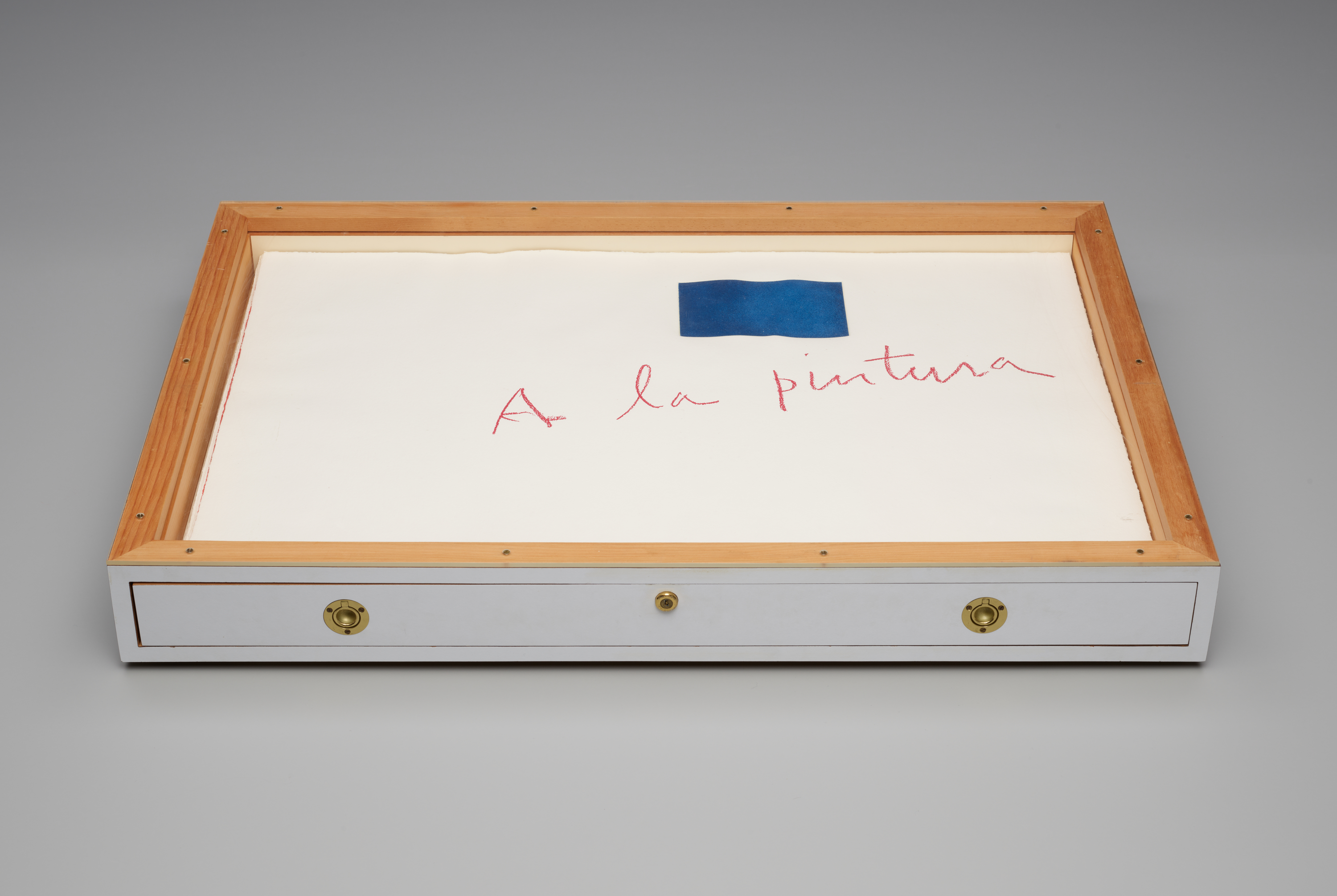 A partially painted wooden box containing the sheets of “A la pintura” against a gray ground. The box has an open wood face and a painted white side with brass hardware for sliding out the drawer containing all of the sheets of paper.