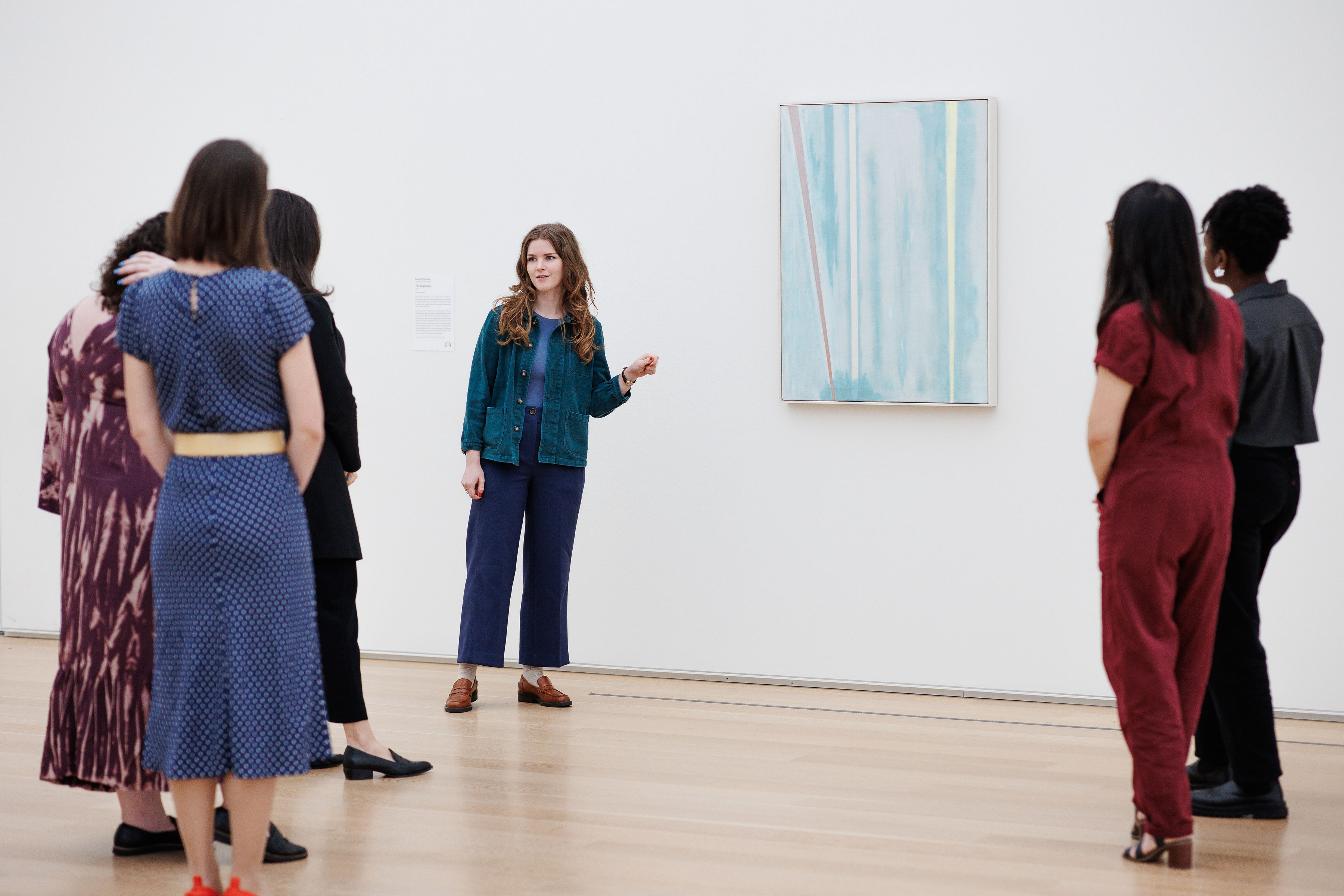 Photo of a group of young women in a white art gallery, seen from the back, viewing Barnett Newman's painting A Beginning, an abstract work featuring a light-blue mottled background and sharp, vertical lines in pink, pale yellow, and white, the pink one on a slight diagonal. A young woman with light skin and light-brown curled hair stands before the artwork, gesturing as though speaking about it.
