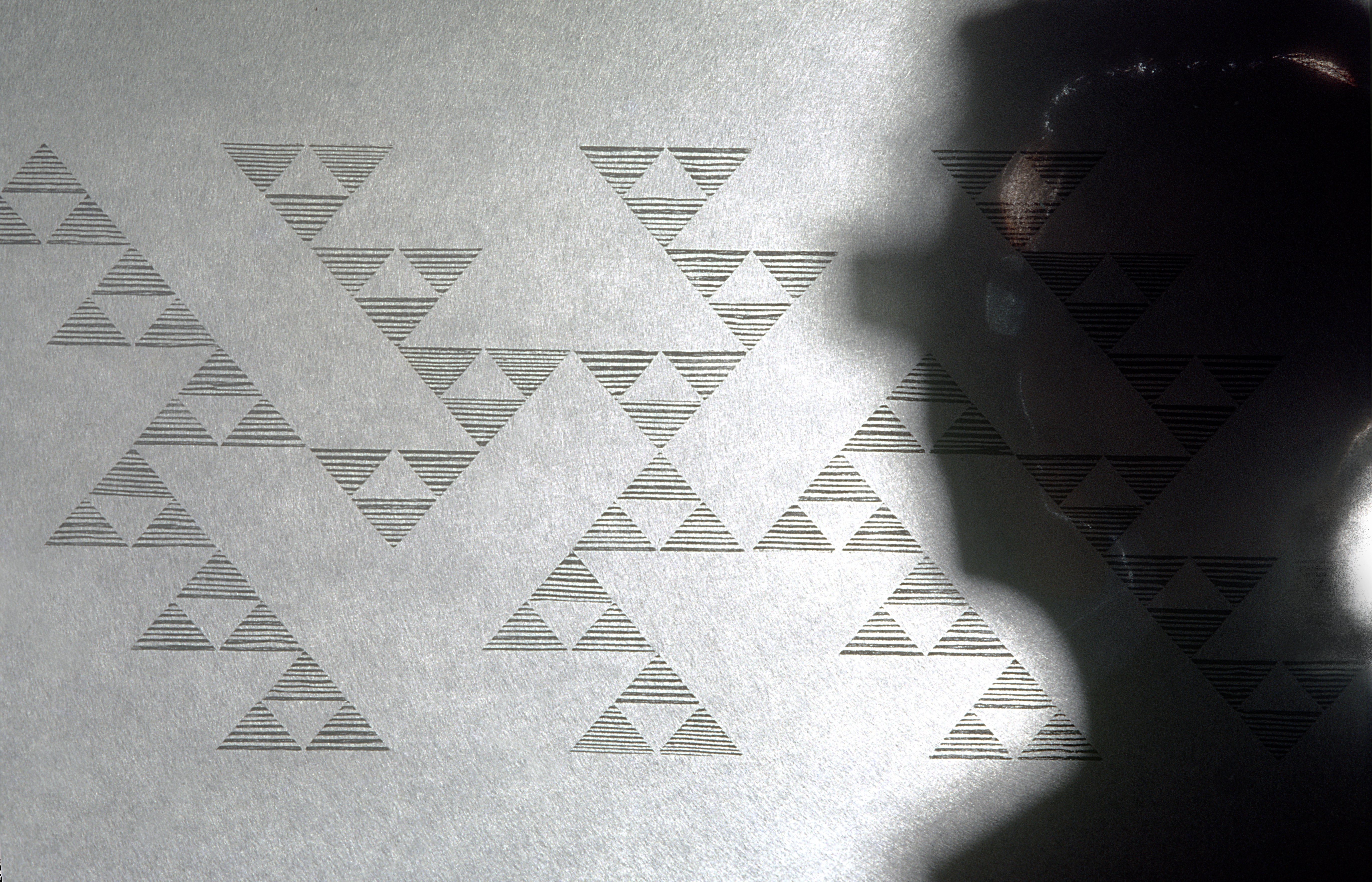 In a black-and-white photograph, pyramid arrangements made of smaller triangles form a contiguous trail across a graywall. The triangles that compose the pyramid are made of uneven, horizontal stripes. On the right a shadow of a woman's facial profile looks onto the arrangement. The woman wears glasses.