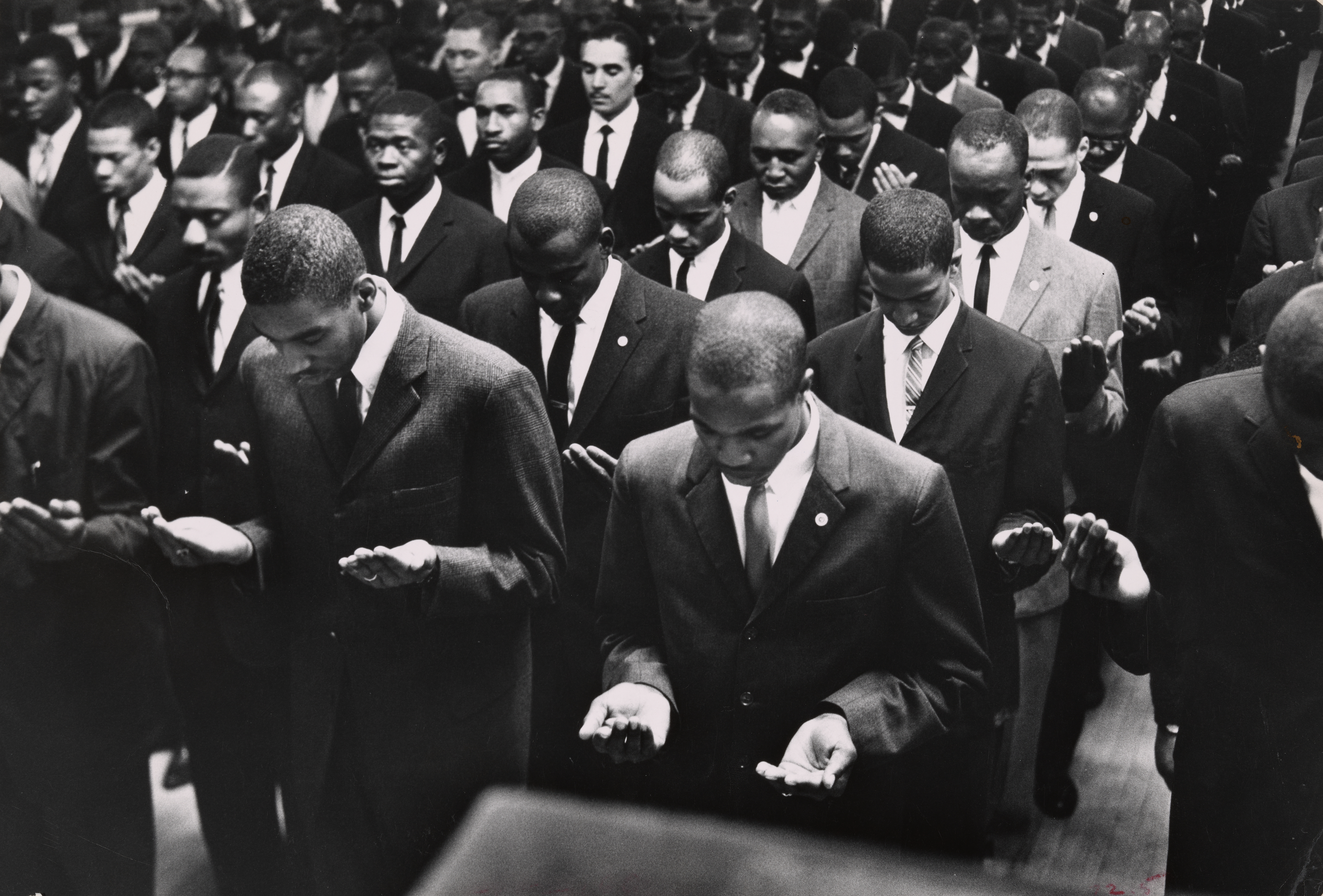 A large group of men dressed in dark suits and ties stand closely packed in rows, facing forward with heads bowed and palms raised in front of them in a gesture of prayer or solemn reflection. Their expressions are serious and focused, and the tightly framed composition emphasizes unity and discipline. The setting appears to be an indoor gathering space with a subdued, formal atmosphere.