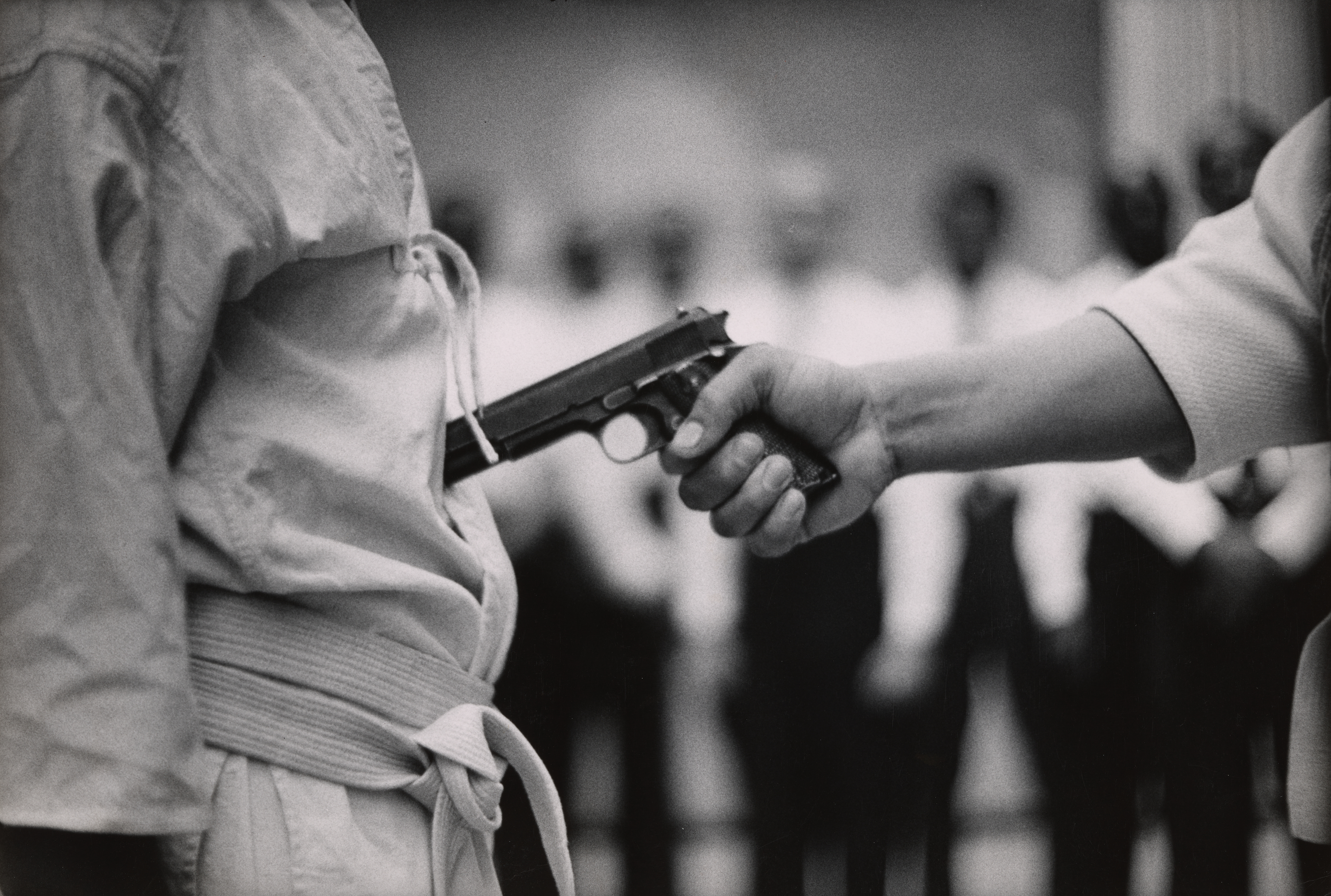 Close-up of a person in a martial arts uniform standing still while another person presses the barrel of a handgun into their abdomen. The handgun is held firmly at close range, with the person’s arm extended from the right side of the frame. In the background, a blurred group of people in uniform appears to be observing, suggesting a demonstration or training scenario.
