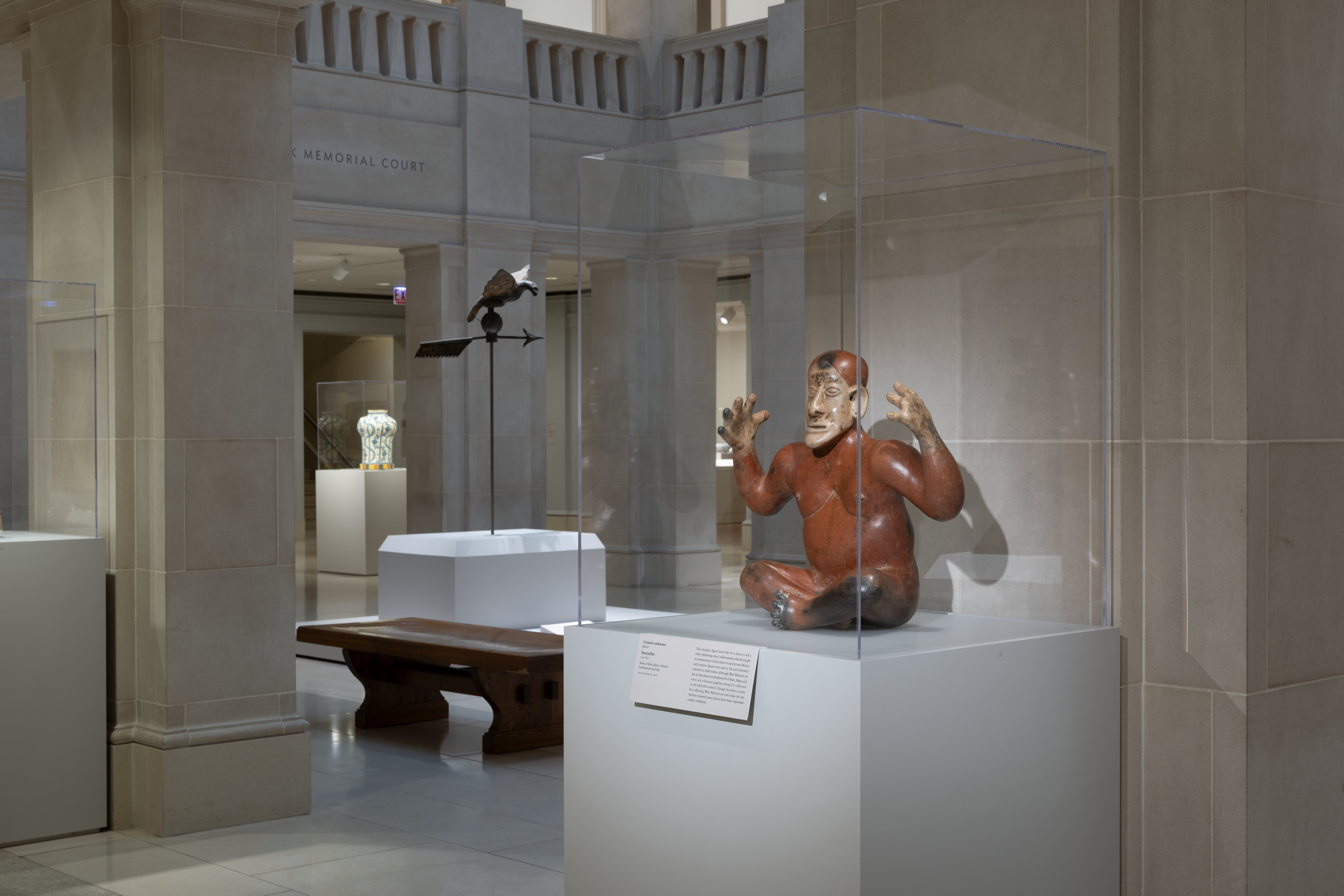 A color photograph of a museum gallery shows a large reddish-brown seated human figure sculpture in a clear case at right, surrounded by beige stone columns, while at left and center are a dark wood bench, a black metal bird sculpture on a stand, and white display pedestals.