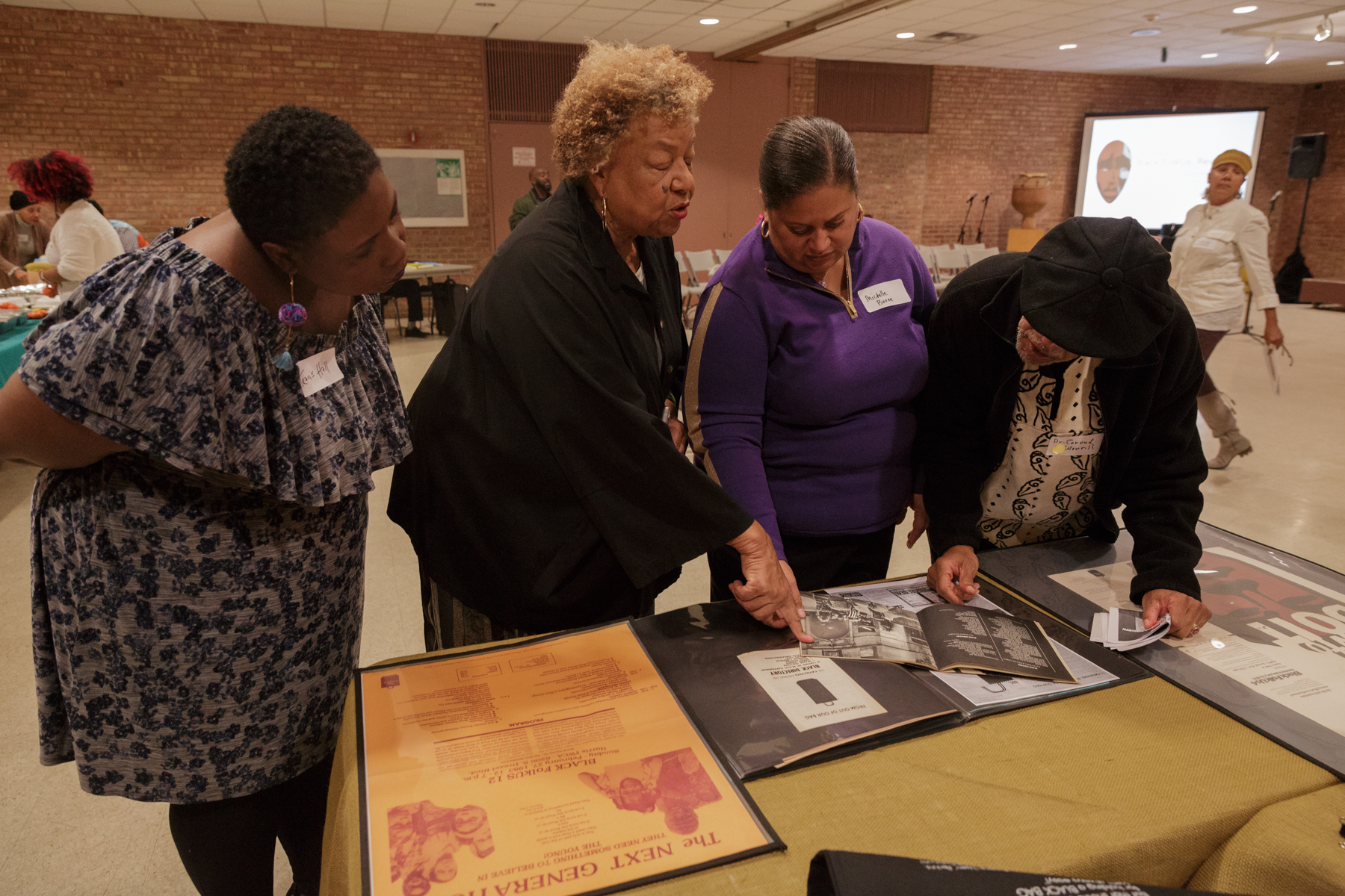 Color photograph of four adults gathered around a table, closely examining documents and posters laid out under clear coverings. The woman in the center, with light brown hair and wearing a black top, gestures toward a page in an open binder as the others lean in attentively. The table displays large printed materials including a bright orange poster and black-and-white text documents. All four people wear name tags. In the background, a person walks past a projection screen and folding chairs in a large room with brick walls and a tiled ceiling.