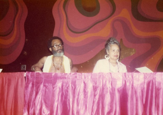 Color photograph of two people seated behind a table draped with a pink satin cloth. The man on the left is shirtless, wearing large glasses, multiple beaded necklaces, and a white cloth draped over his shoulders. The woman on the right wears a light pink top and large hoop earrings, and has gray hair styled in an updo. Both appear to be reading from or seated before papers. The background wall features a bold, abstract pattern of swirling pink, orange, and black shapes.