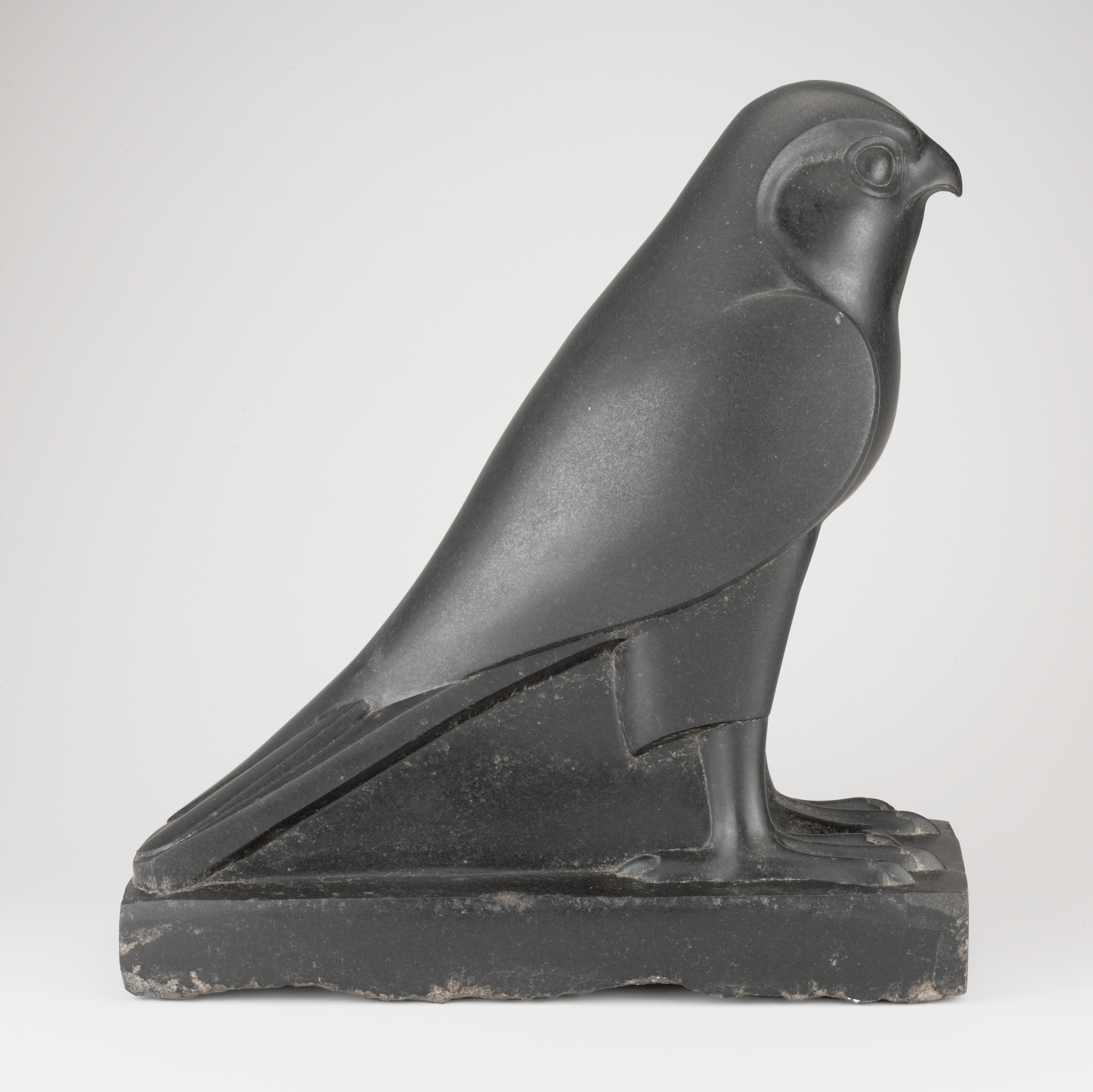A dark gray bird sculpture is shown in profile facing right. Starting at the left, a long tapered tail leads forward past folded wings to a rounded body and a head with a short, hooked beak and a round, raised eye at the far right. At the bottom foreground, clawed feet stand on a thick dark-gray rectangular base, set against a plain white background.