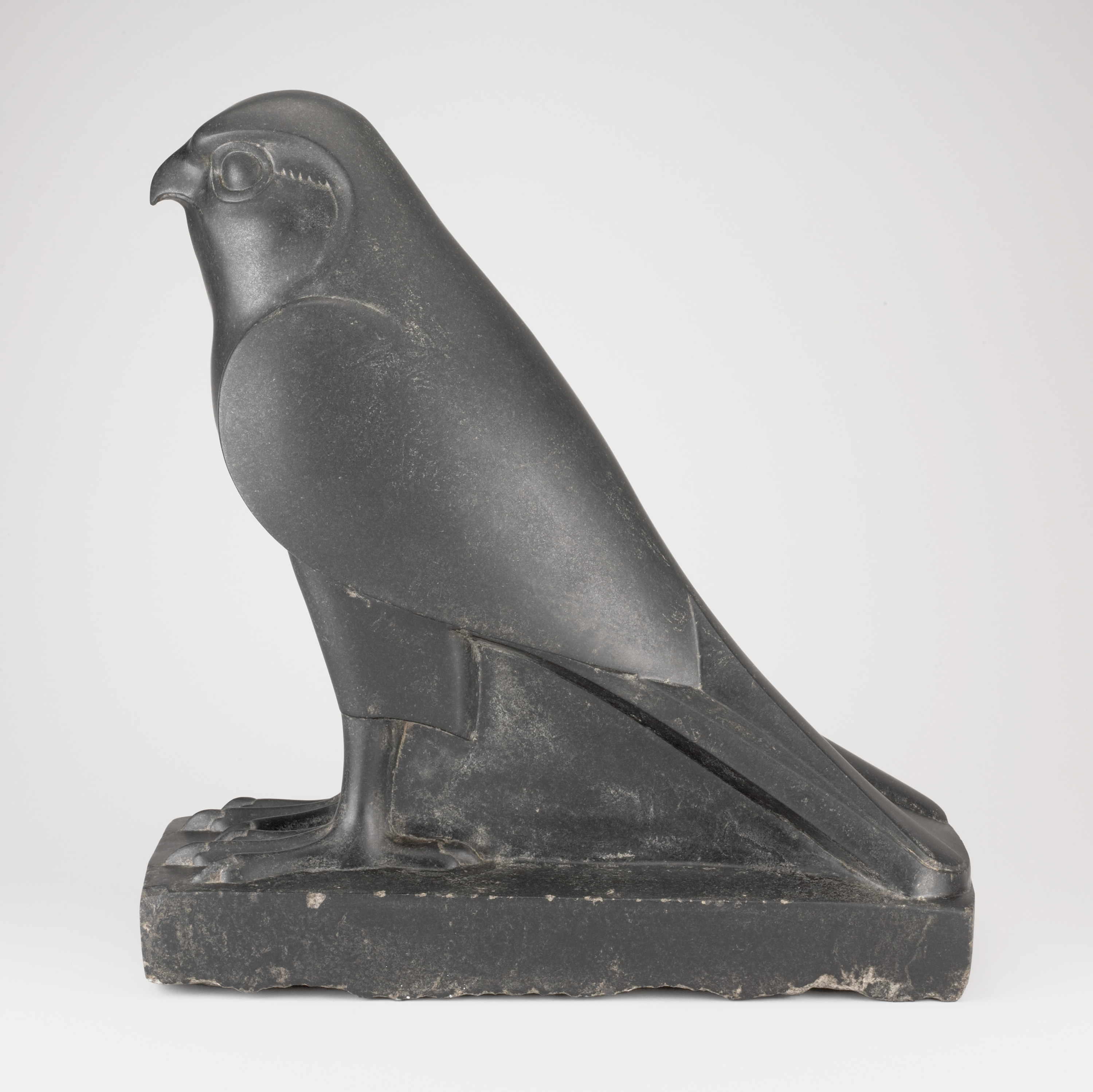 A dark gray bird figure is shown in left-facing profile, with a rounded head, a curved beak, and a circular eye near the top. Moving downward, smooth folded wings meet a compact body; at the bottom, the feet with four toes rest on a rectangular base, and a long tail extends along the back of the base. The figure is uniformly dark gray against a plain white background, and the bird’s body is much taller than the base is thick.