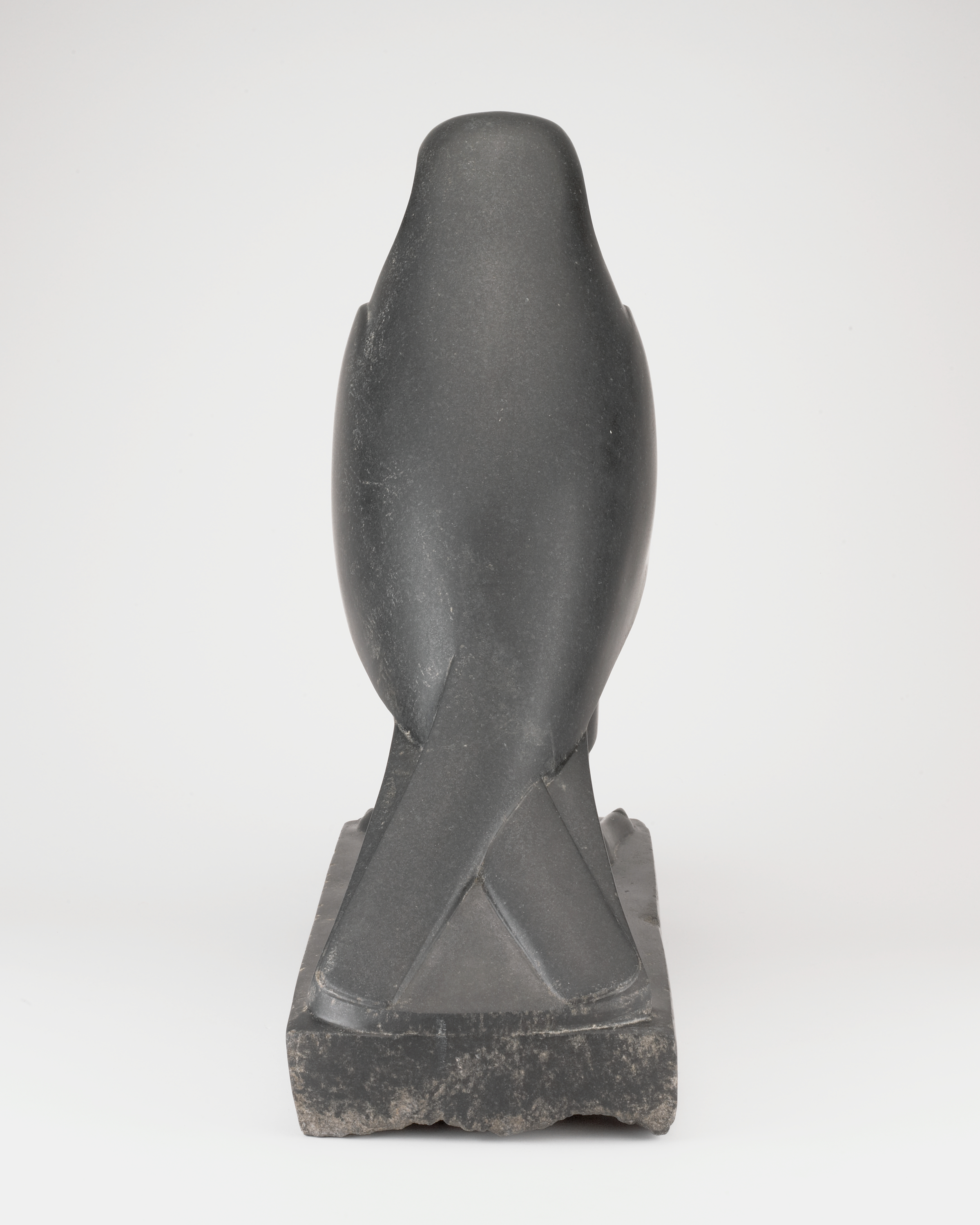 From top to bottom: a smooth, rounded bird-like head seen from behind, followed by an oval torso that occupies most of the height. At the bottom, two crossed, angular forms meet a rectangular platform. The figure is dark gray with slightly lighter wear along the lower edge, and the body appears larger than the narrow base against a plain white background.