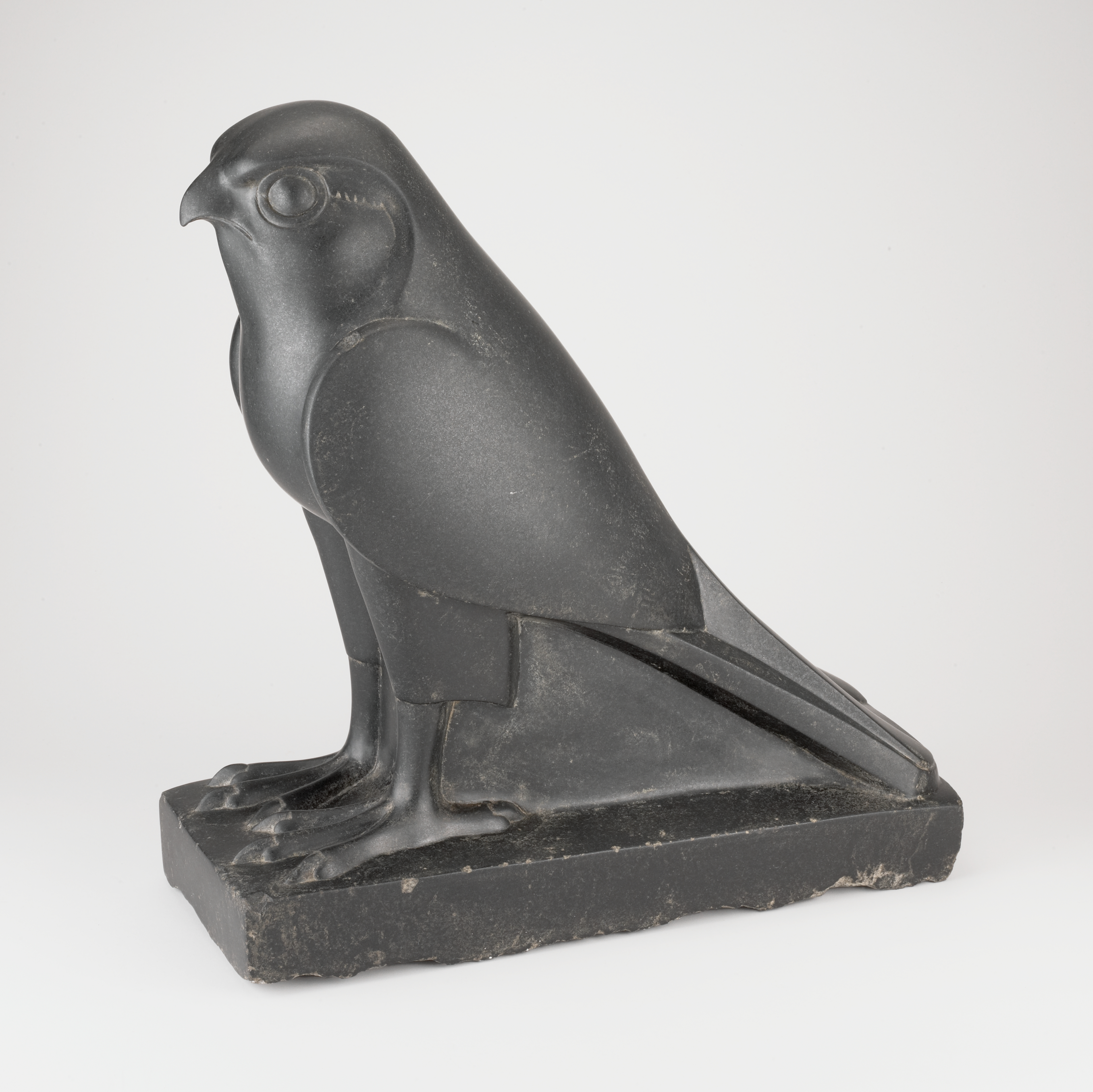 At left, a dark gray-black bird of prey faces left with a rounded head, hooked beak, and a circular eye. Moving right, smooth folded wings lie along its sides and a long, tapering tail extends toward the back right. In the foreground, the bird stands on a low rectangular base with defined talons at the front; the background is plain white.