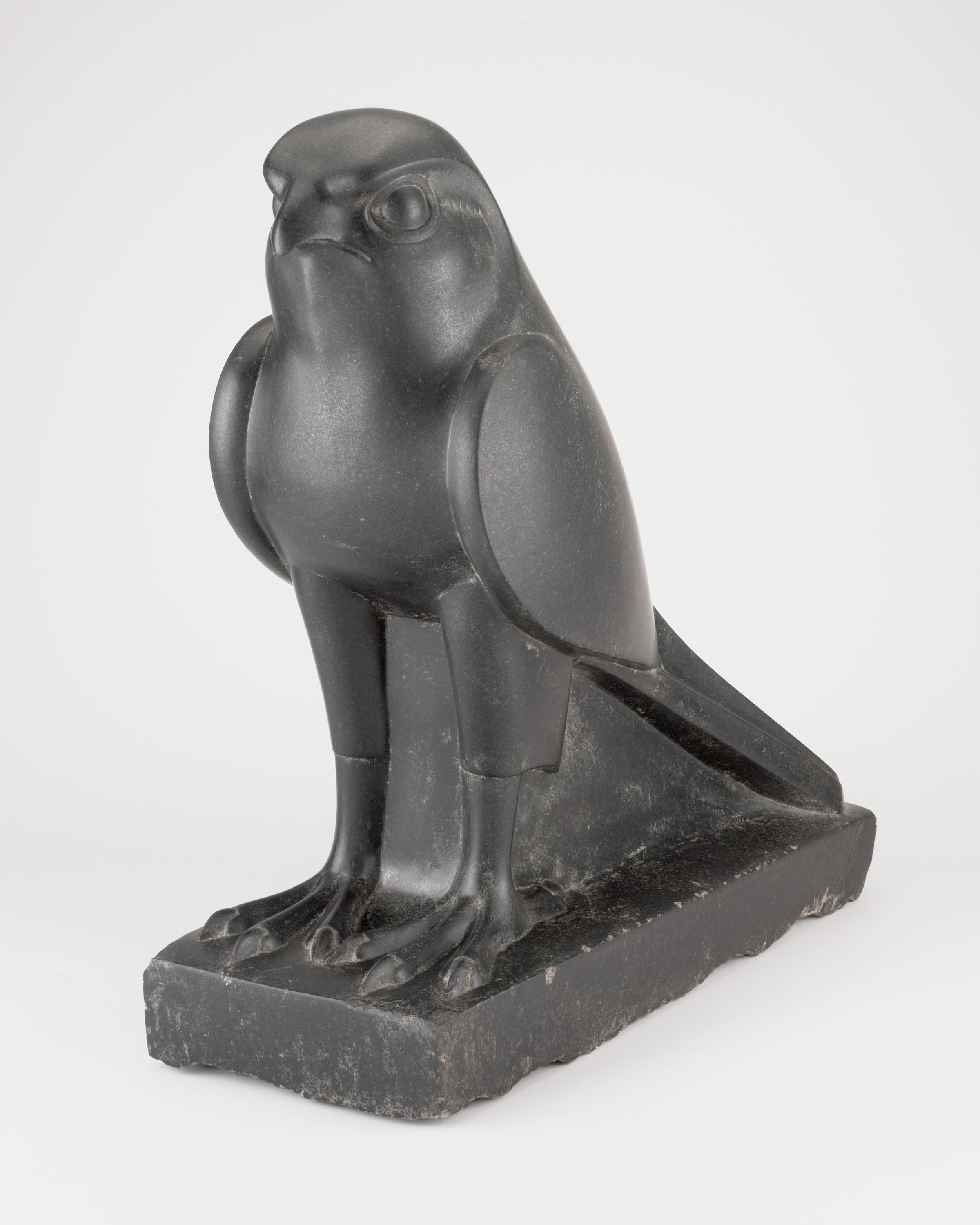 At the top, a stylized dark gray bird head faces forward with a short curved beak and round, raised eyes. Below, smooth oval wings rest against a broad chest, and two straight legs end in four clawed toes on each foot. The figure stands on a rectangular base with worn, lighter gray edges, and a long flat tail extends backward along the base. The background is plain white.