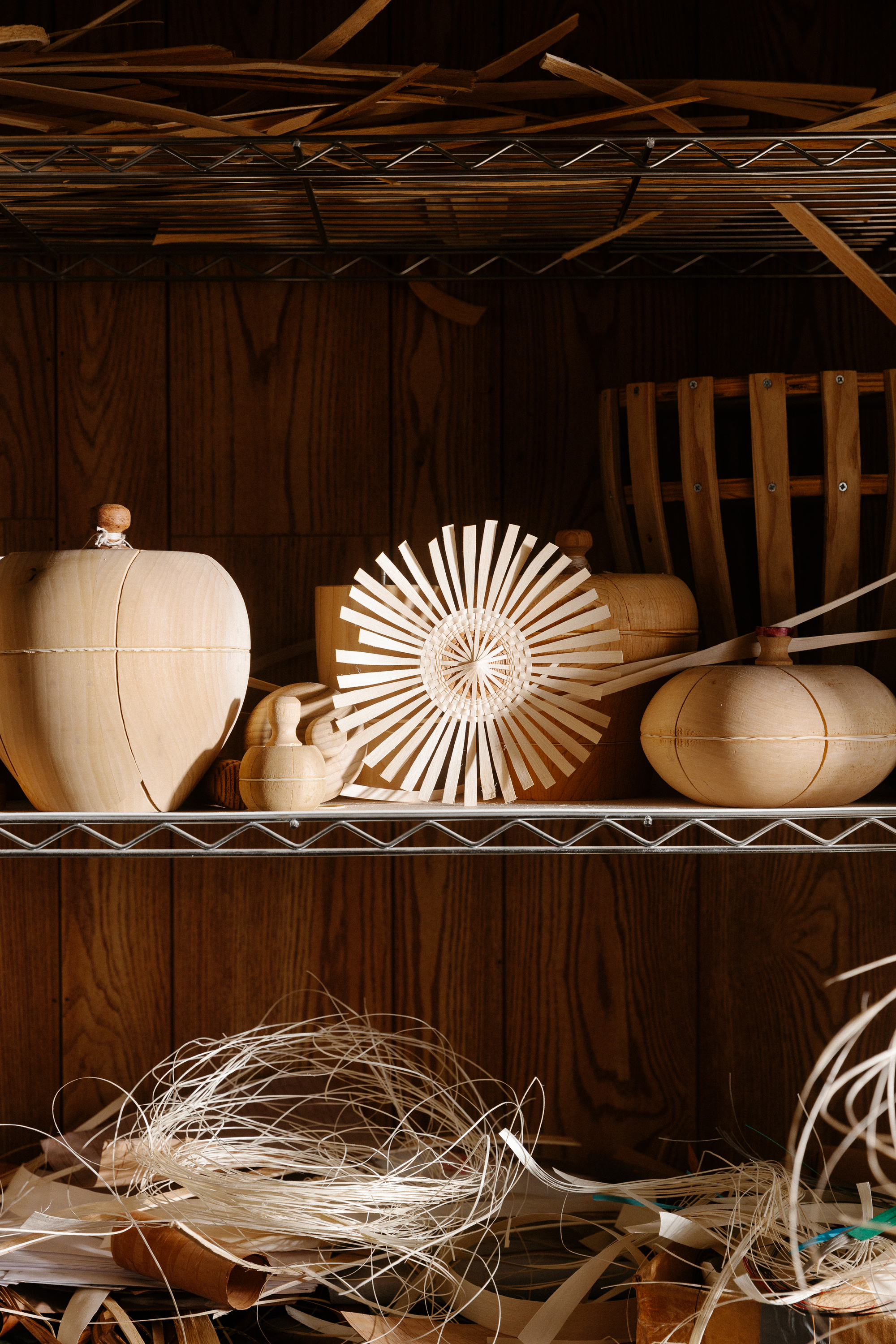 A color photo of two shelves. On the bottom are a few tangles of thin strips of blond wood. On the top are solid vessel-shaped pieces of wood and a sunburst-like design composed of thin wood strips.