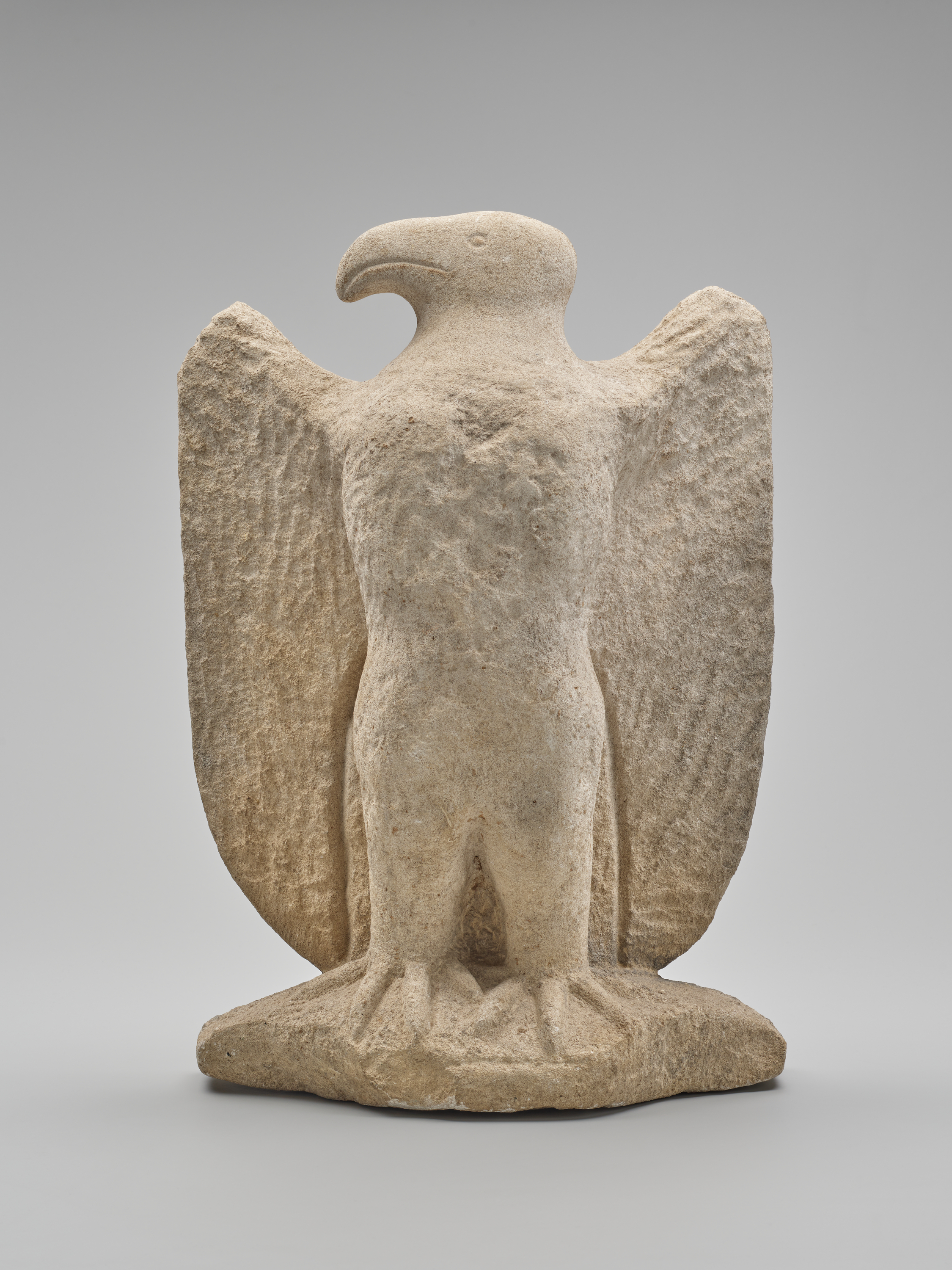 A beige stone sculpture of an eagle stands at center on a rough base, its curved beak facing right, taloned feet at bottom center, and broad wings stretching to left and right, all with a textured surface against a plain gray background.