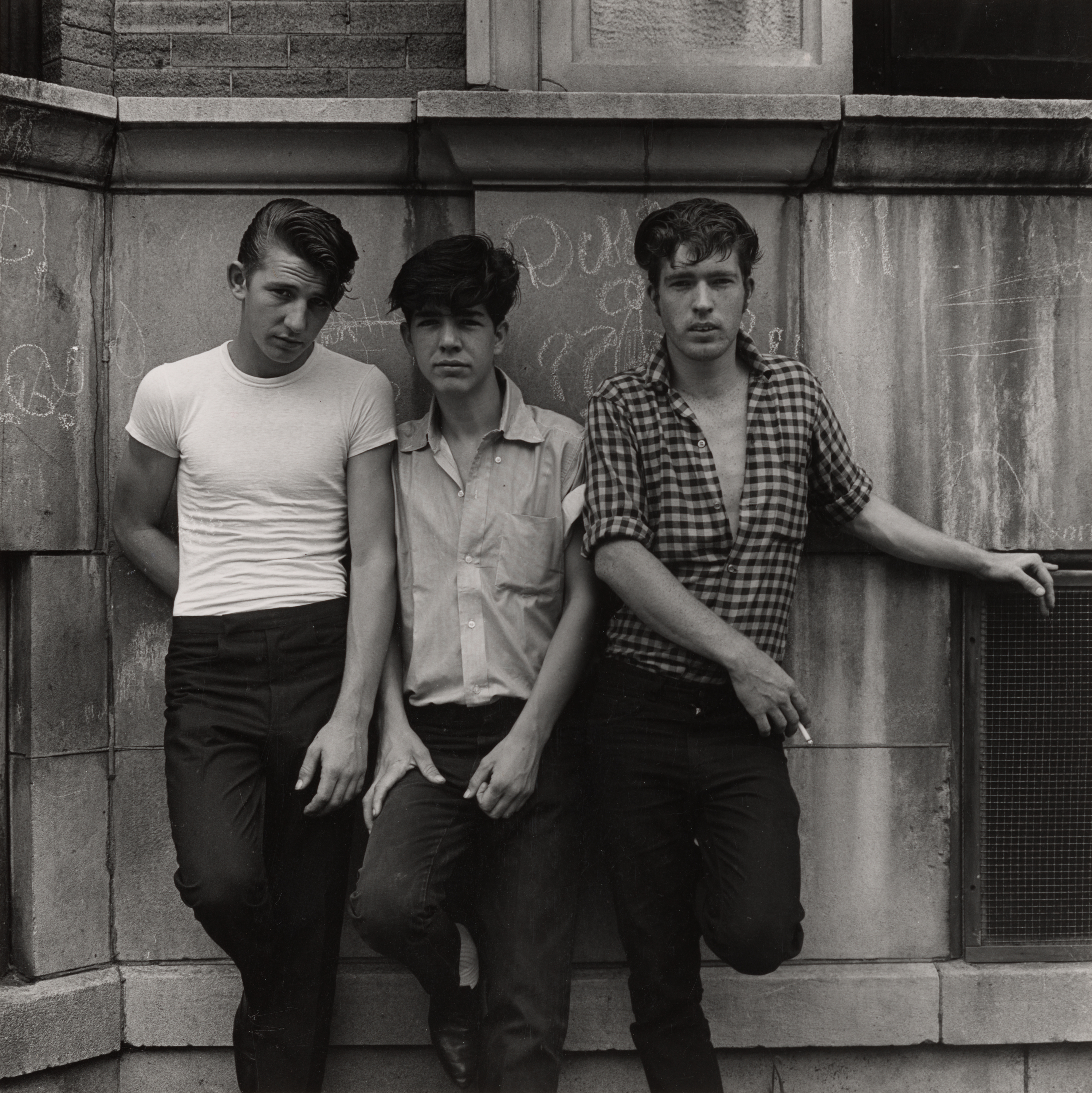 Three teenage boys stand close together against a stone wall marked with chalk graffiti. The boy on the left leans with his hands in his pockets, wearing a tight white T-shirt and dark pants. The boy in the center wears a short-sleeved button-down shirt and rests one hand in his pocket. The boy on the right wears a checkered shirt open at the collar and holds a cigarette in one hand, with his other arm extended to the side. Their expressions are serious and direct, conveying a sense of quiet confidence and camaraderie.