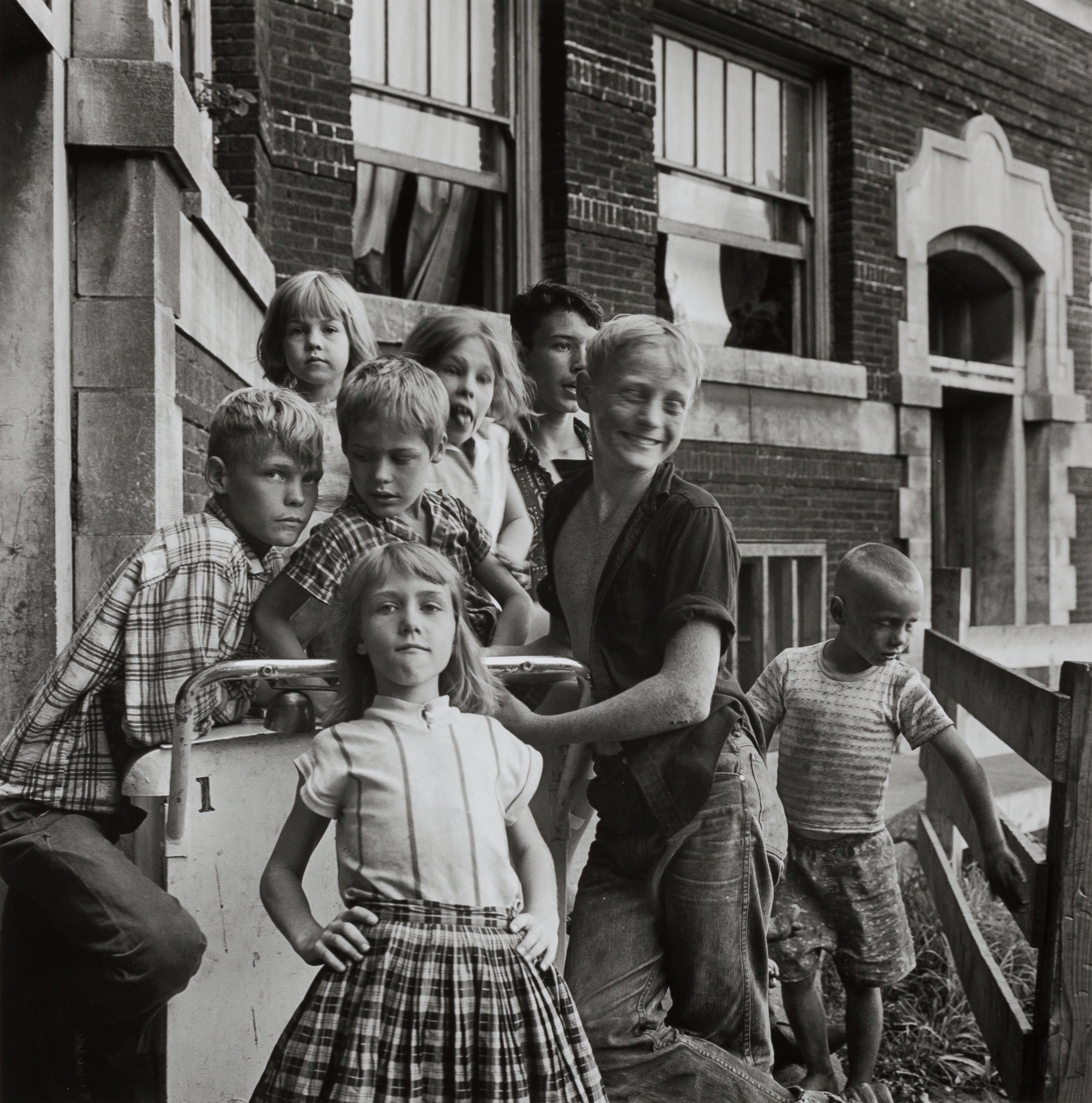 A group of nine children gather on a sidewalk beside a brick apartment building. In the foreground, a girl stands confidently with hands on hips, wearing a plaid skirt and short-sleeved shirt. Behind her, boys and girls lean on a metal railing and stand on stairs, some facing the camera while others turn to each other, talking or laughing. The children wear casual clothes, and the building's stone and brick façade, along with open windows and curtains, forms the backdrop.