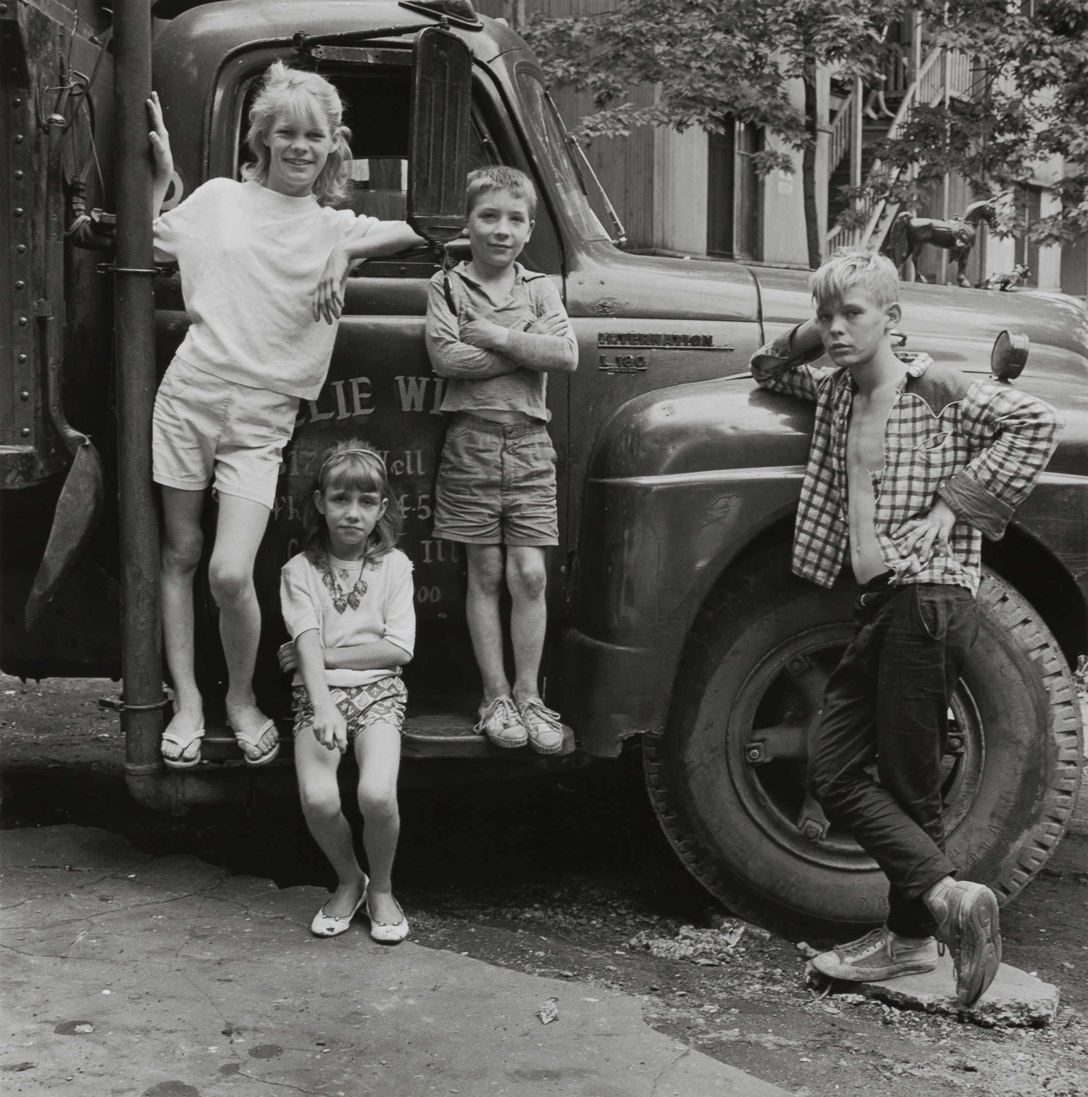 Four children pose in front of a parked delivery truck. A girl and boy stand on the truck’s running board, smiling with arms crossed. A younger girl sits below them with arms folded and a serious expression. Another boy leans against the front fender, shirt open and gaze directed away. The truck has painted lettering on the door, and stairs and balconies are visible in the background. The children wear casual warm-weather clothing.