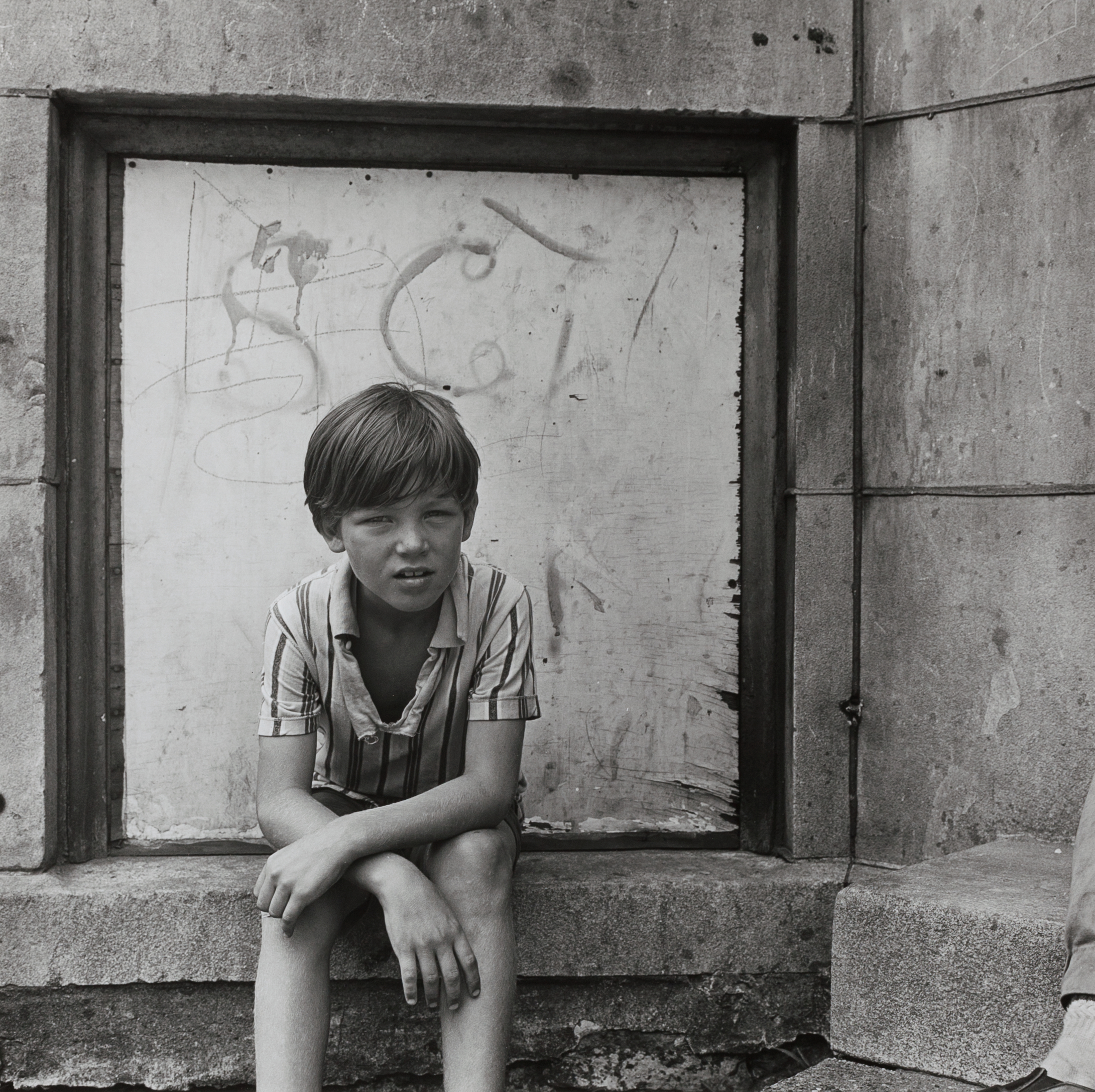 A boy with tousled hair sits on a concrete ledge with his legs bent and arms resting on his knees. He wears a short-sleeved, striped button-up shirt and shorts. His expression is squinting and serious, directed slightly toward the camera. Behind him is a worn, boarded-up window set in a stained stone wall, marked with faint graffiti.
