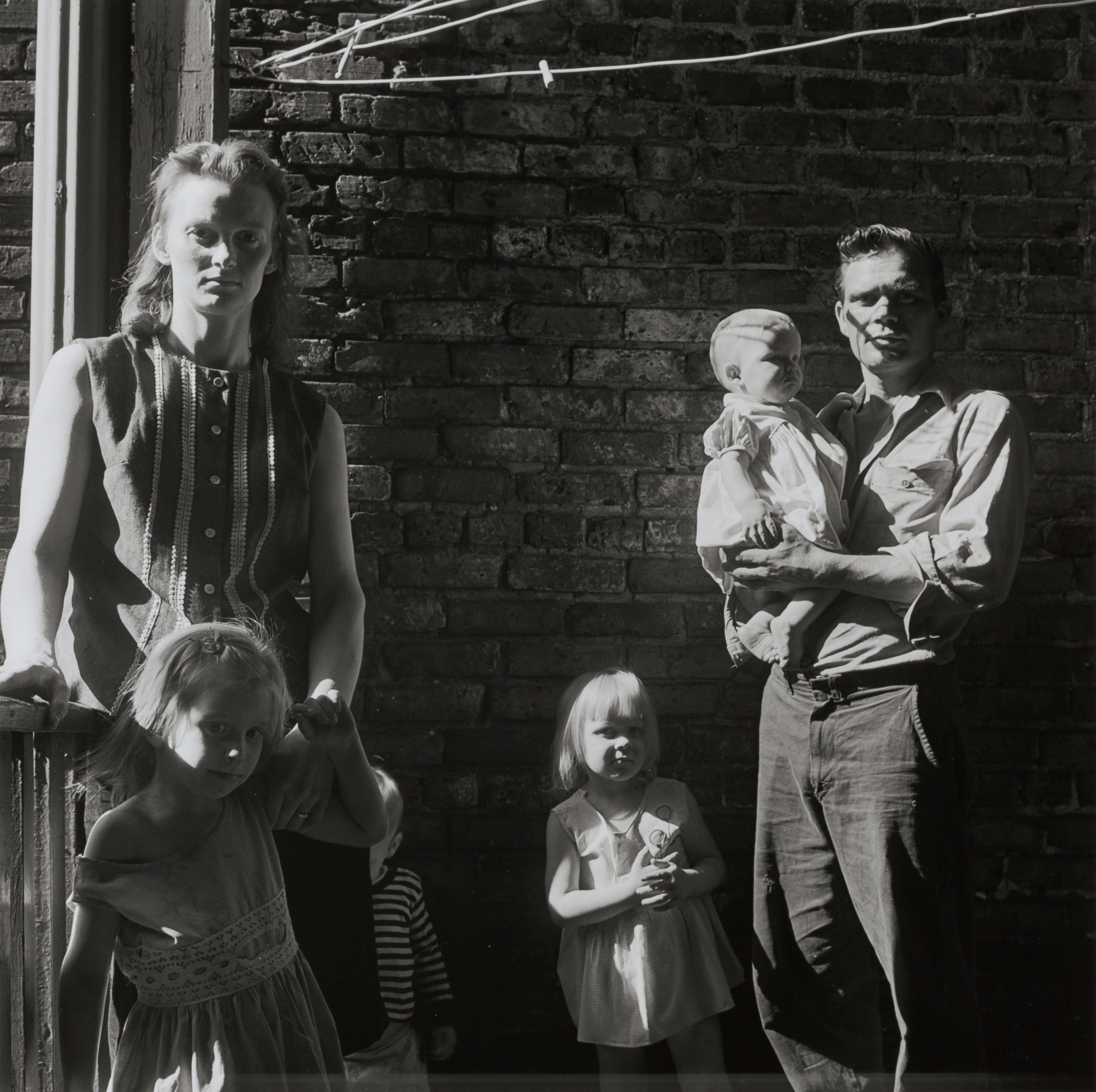 A family of six stands in partial sunlight against a weathered brick wall. On the left, a woman in a sleeveless button-down top rests her hand on a railing, with a young girl in a dress leaning against her. Two other children stand near the center—one in a striped shirt partially hidden, the other in a sleeveless dress holding her hands together. On the right, a man in rolled-up sleeves holds a baby in his arms. Harsh shadows cross their faces, and a clothesline stretches above, contributing to a stark and intimate scene.