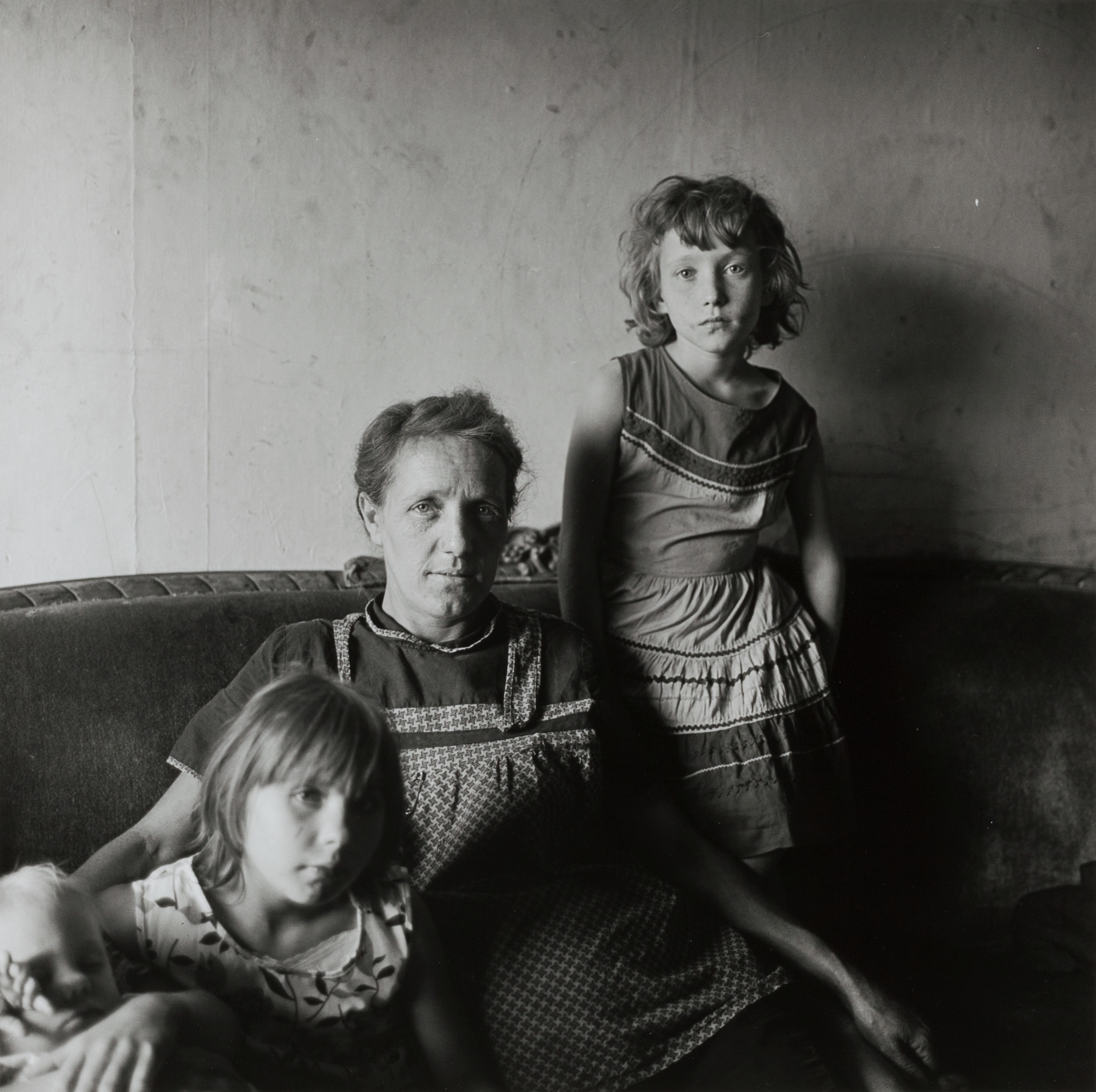 A woman sits on a worn sofa with a baby sleeping in her lap and a young girl leaning beside her, while an older girl stands to her right. All three children have light hair, and the woman wears a patterned apron. They are in a bare room with a plain wall, lit softly from the left, creating strong shadows that emphasize their expressions and the texture of their clothing.