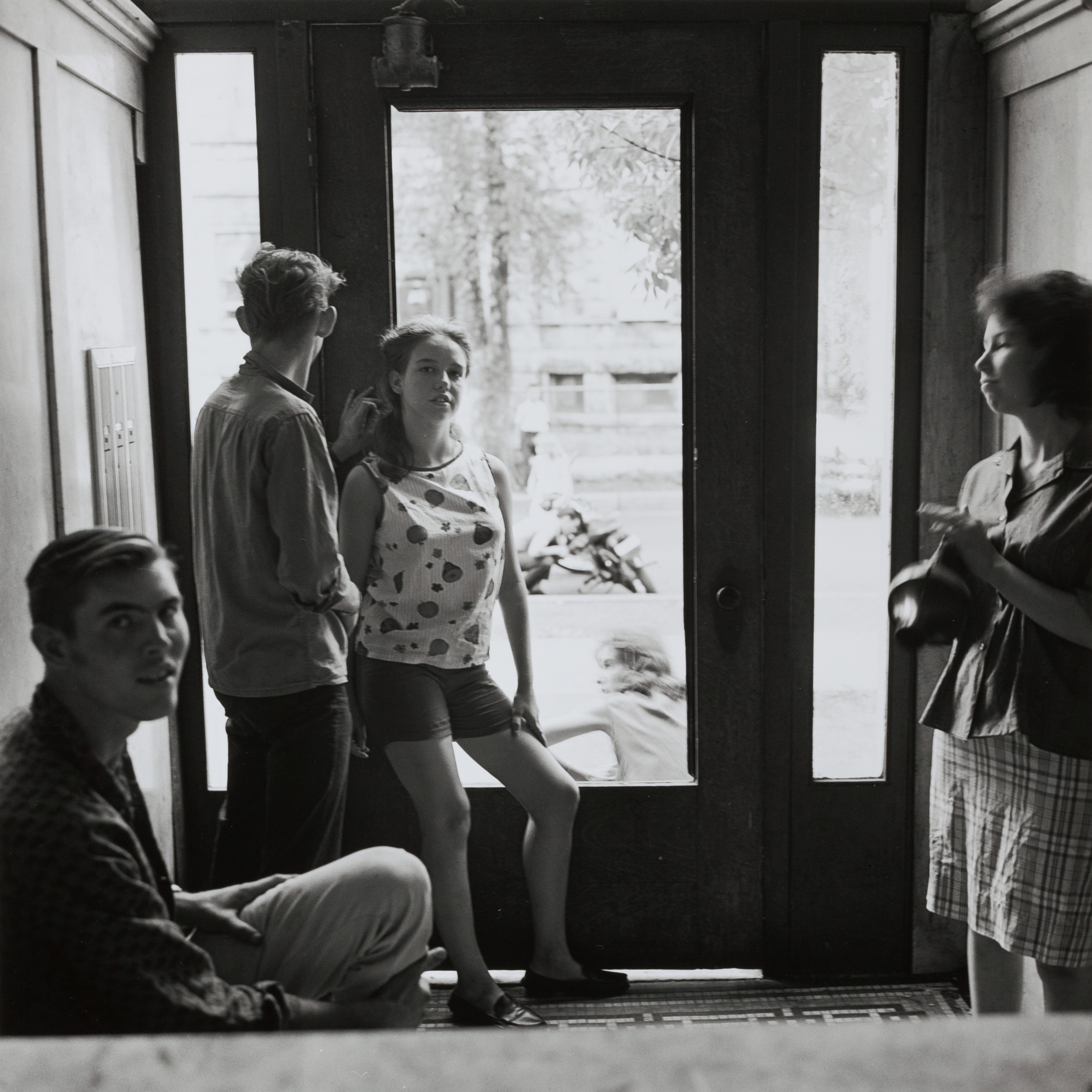Five teenagers gather in the doorway of a building, framed by glass panels that show the street and sidewalk outside. A young man sits on the floor in the foreground, looking toward the camera, while another boy and girl stand near the open door, casually posed. Another girl stands to the right, mid-motion as if adjusting something in her hands. Light from outside softly illuminates their faces, contrasting with the darker interior. The scene captures a quiet, candid moment of adolescent pause and observation.