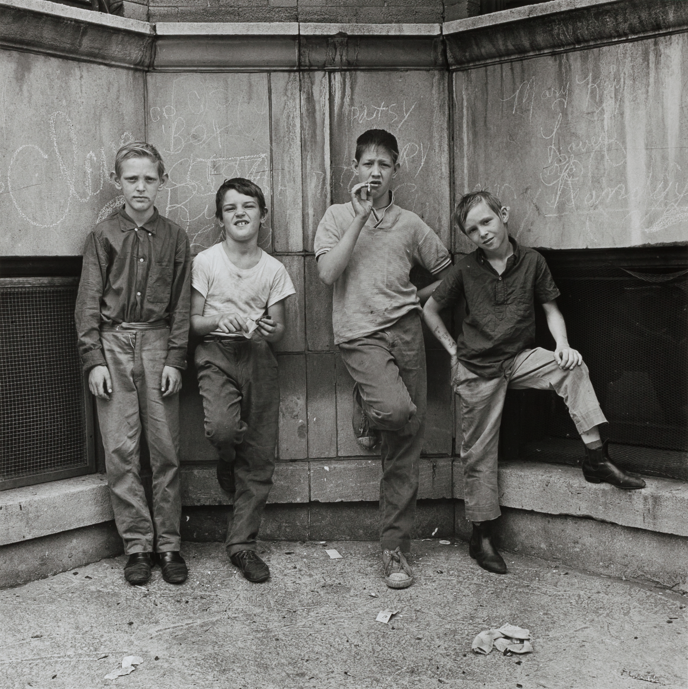 Four boys stand in a row against a stone wall, each with a casual or defiant pose. They wear rumpled shirts and pants, with the second boy from the left gripping a slingshot and the third holding a cigarette. The background wall is covered in chalk graffiti with names like "Patsy" and "Mary Kay." The scene conveys a gritty urban energy, with scattered litter on the pavement and a strong sense of camaraderie among the boys.