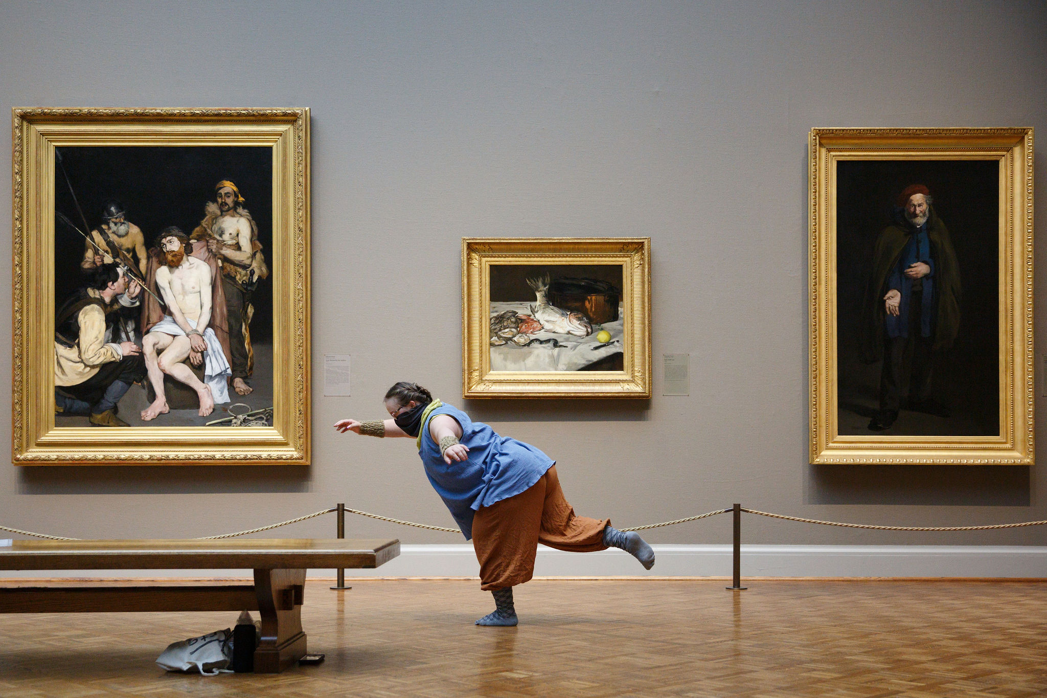 Photograph of a figure in a black face covering performing dance in an art gallery, their body in a leaning arabesque, large oil paintings behind them.