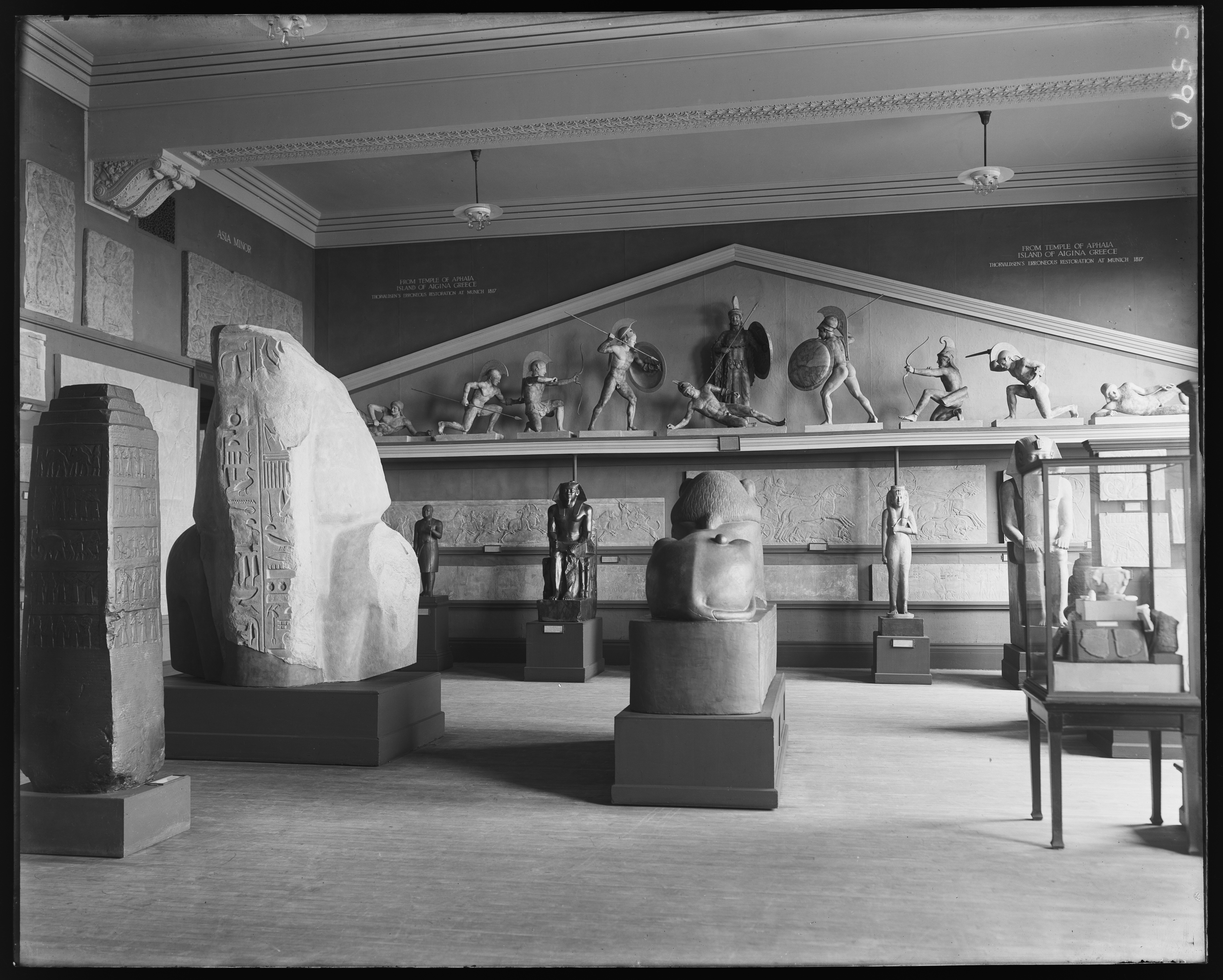 Black-and-white photograph of a museum gallery with warrior figures atop a transom and pedestals on the floor, carved stone-like slabs at left.