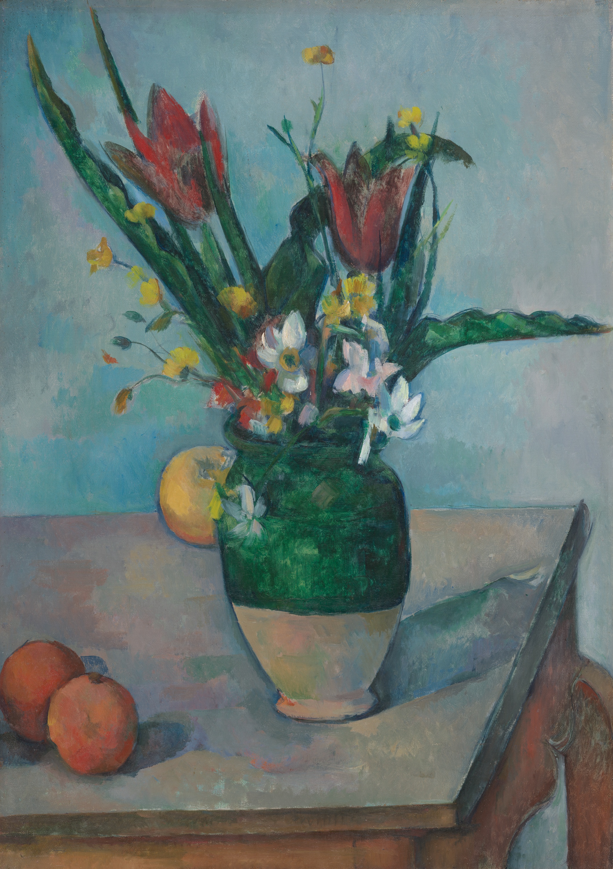 A green-and-cream vase on a light gray tabletop holds tall green leaves that reach toward the upper edge, two large red flowers, and clusters of small white and yellow blossoms, set against a pale blue background. At the lower left, two round orange fruits sit on the table, and a single round yellow fruit rests just behind the vase near the center; the vase is larger than the fruits. Toward the right, the tabletop continues to the corner with a table leg visible, and shadows from the vase and leaves spread across the surface.