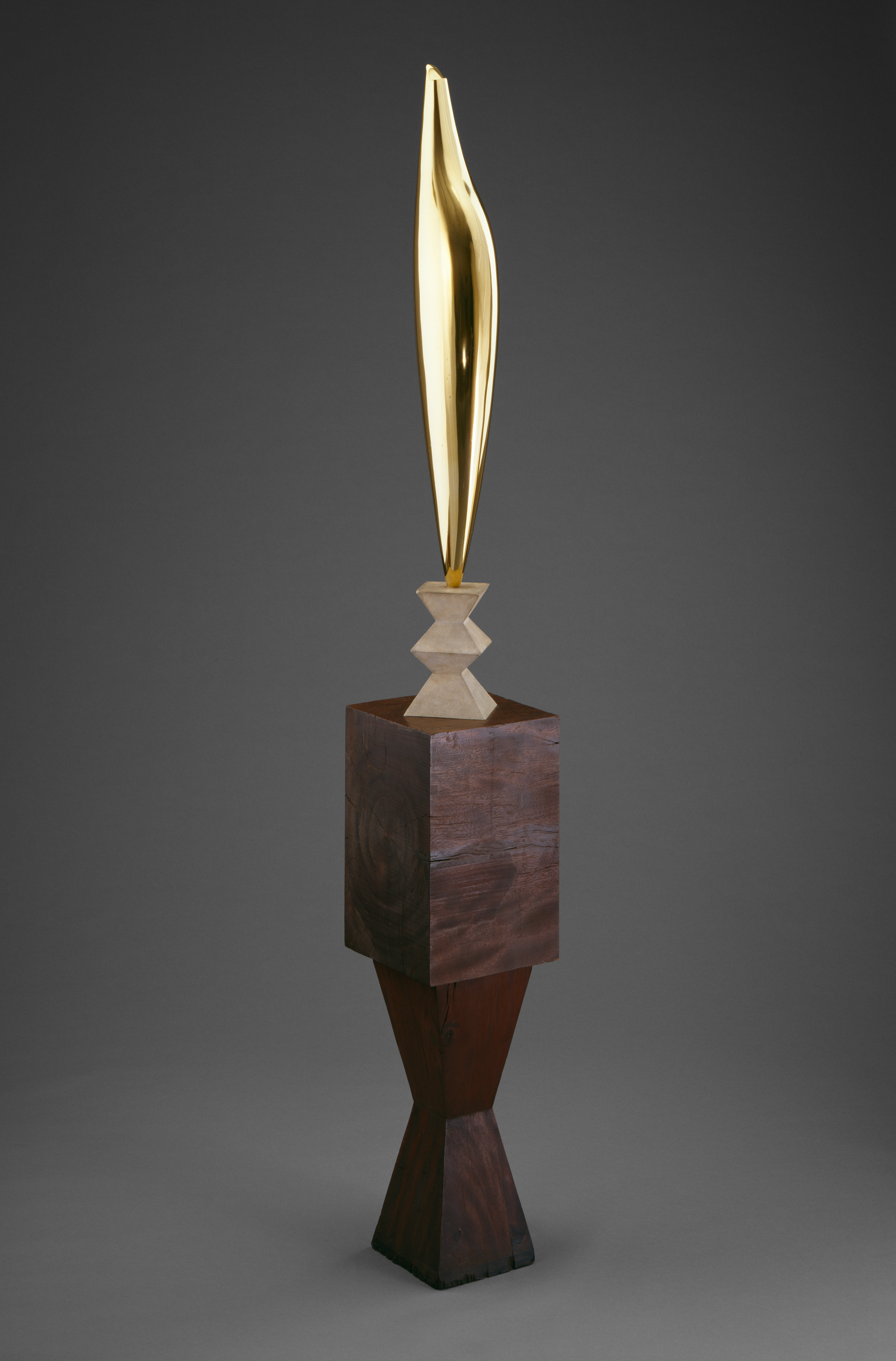A color photograph shows at center a tall, polished gold, leaf-like metal sculpture atop two stacked light stone pyramids, resting on a dark brown wooden block with an hourglass foot, all against a plain gray background.
