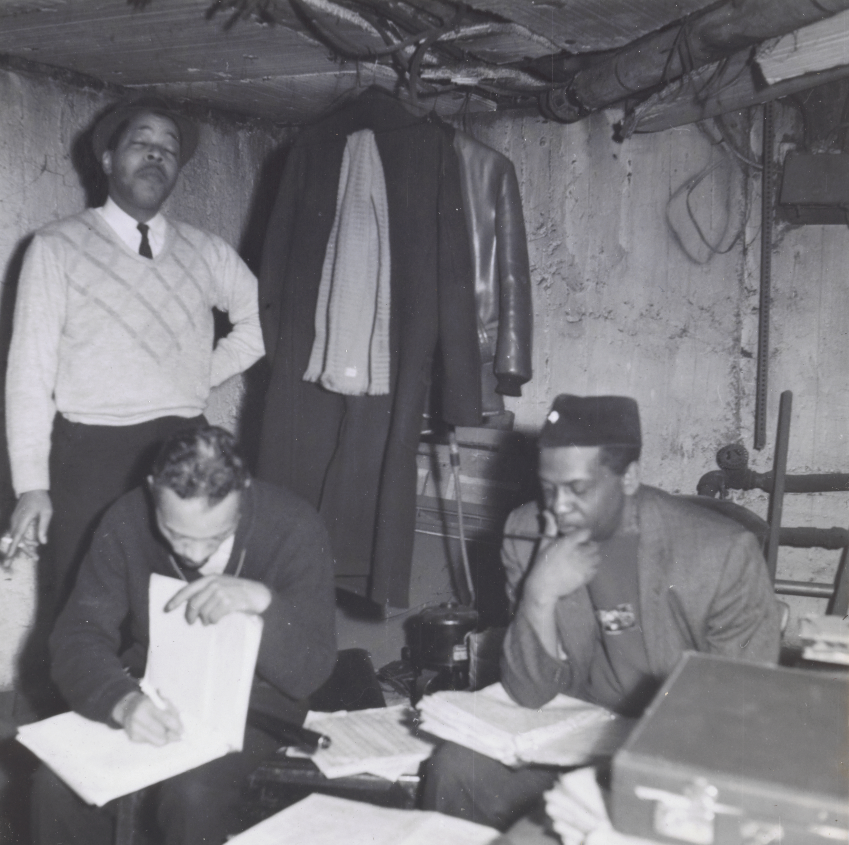 Photograph of three men working in a cluttered basement room with exposed pipes and rough concrete walls. At left, a man in a light sweater and fedora stands with one hand on his hip, holding a cigarette. In the center, a seated man writes on a sheet of paper, leaning over a binder. At right, another man in a fez-style hat rests his chin on one hand while looking down at a stack of papers. Coats and a scarf hang from the ceiling above them, and boxes and documents cover the table in front of them.
