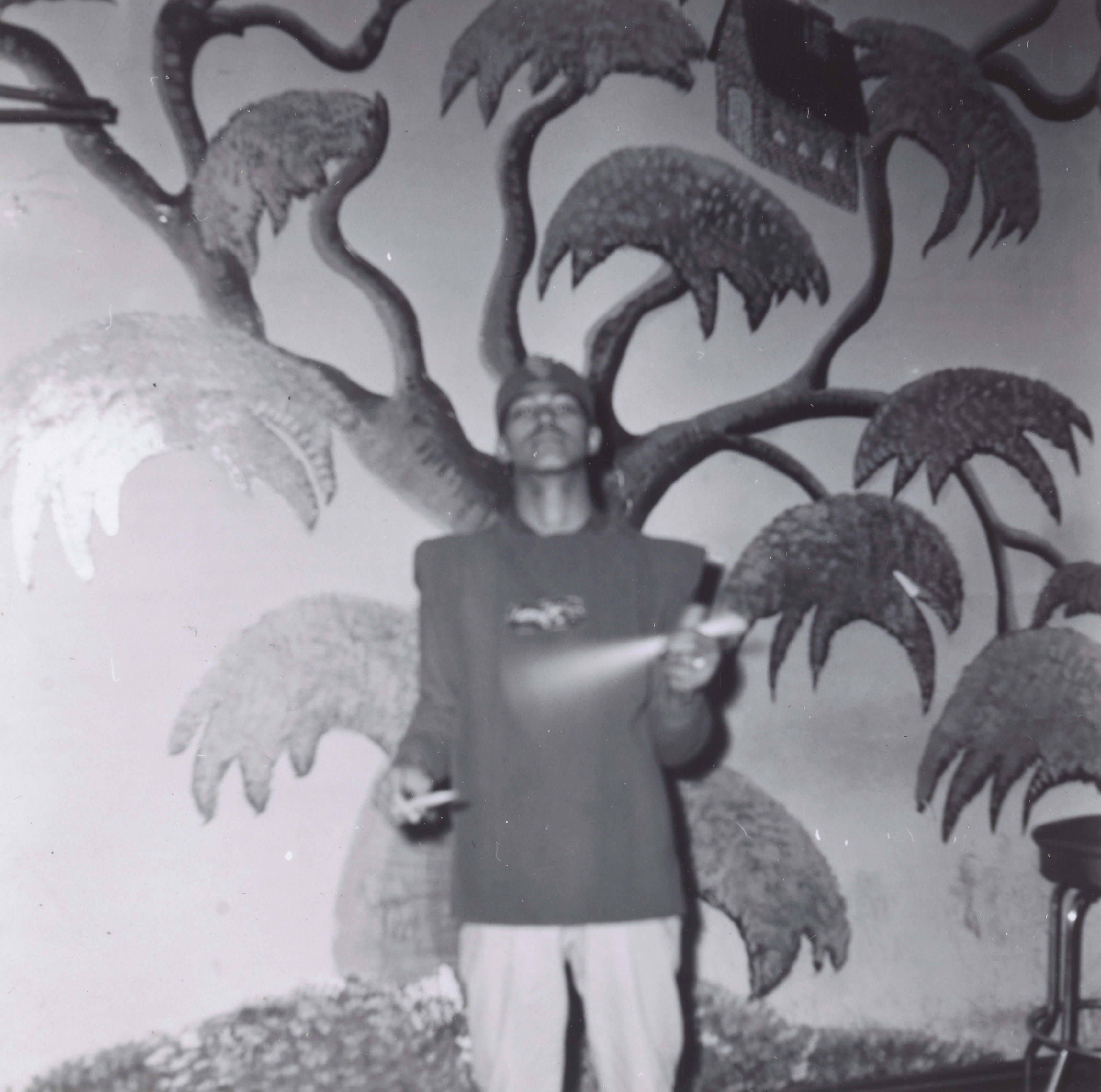 Photograph of a man standing in front of a sculpted wall relief featuring a large, stylized tree with textured leaves and a small house perched in the upper branches. He wears a fez-style hat, dark long-sleeved shirt, and light pants. He holds two drumsticks mid-motion, one of them blurred, suggesting he may be playing or demonstrating a rhythm. A stool is partially visible at the far right.