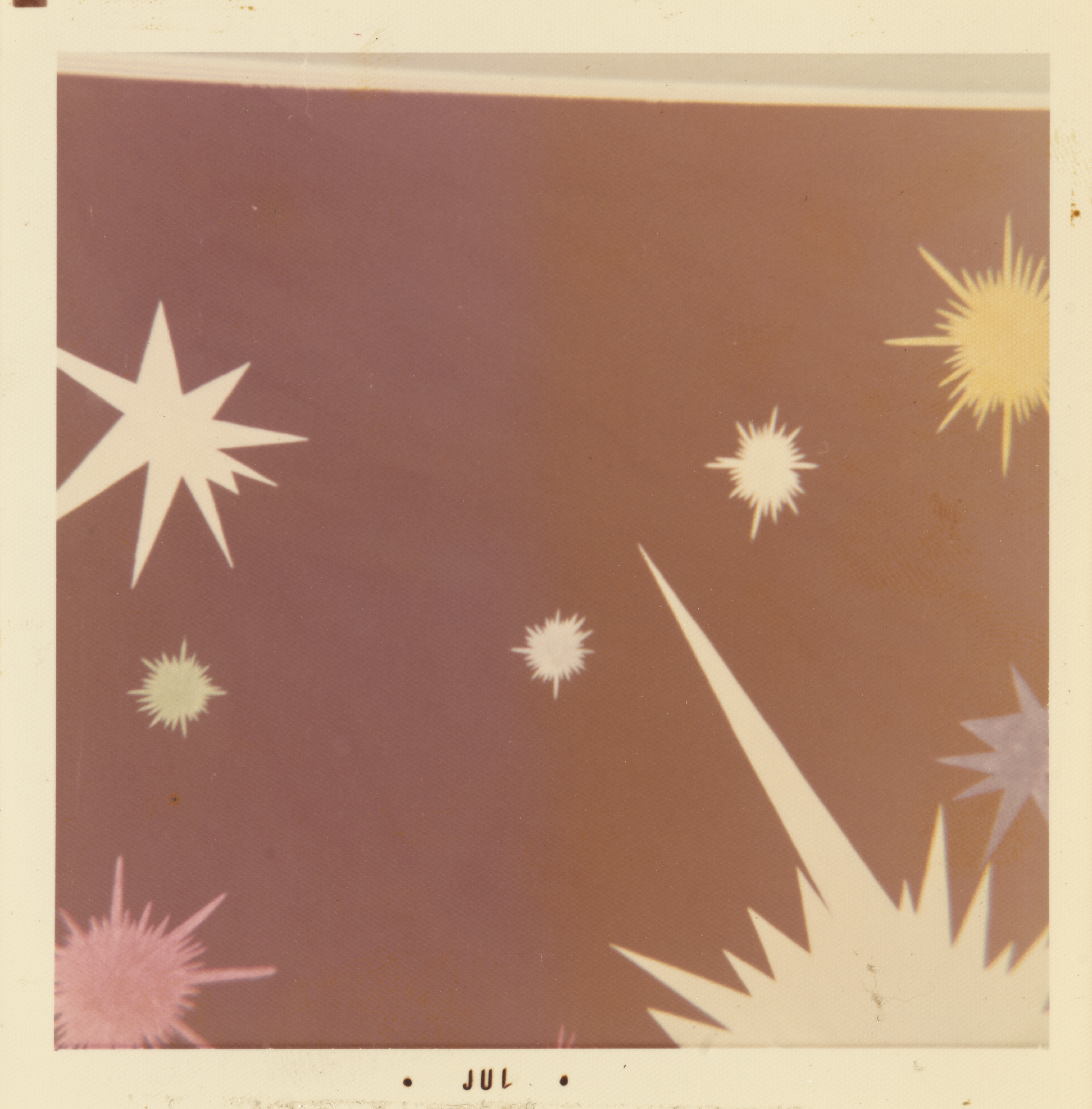 Photograph of a painted mural with a celestial theme on a mauve background. The composition features multiple starburst shapes in white, yellow, pink, green, and lavender, scattered across the frame. A large white burst with long, sharp rays extends upward from the bottom right corner. The top edge shows a narrow strip of pale molding or ceiling detail. The white border of the print includes the text “• JUL •” centered along the bottom edge.