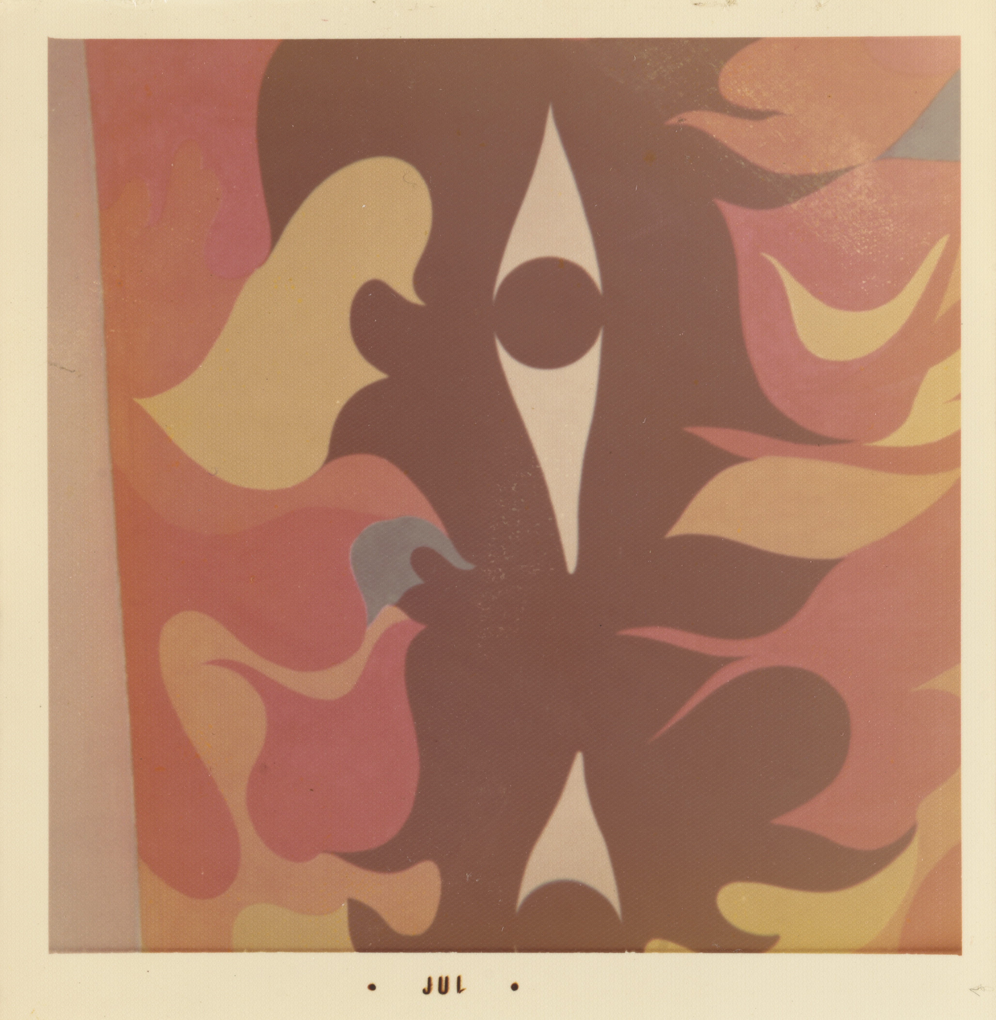 Photograph of an abstract mural featuring a symmetrical composition of curving, flame-like forms in shades of brown, red, pink, yellow, and orange. At the center, a vertical brown shape encloses two white eye-shaped forms, each with a dark circular center. The surrounding shapes radiate outward in soft, flowing layers. The white border of the print includes the text “• JUL •” centered along the bottom edge.