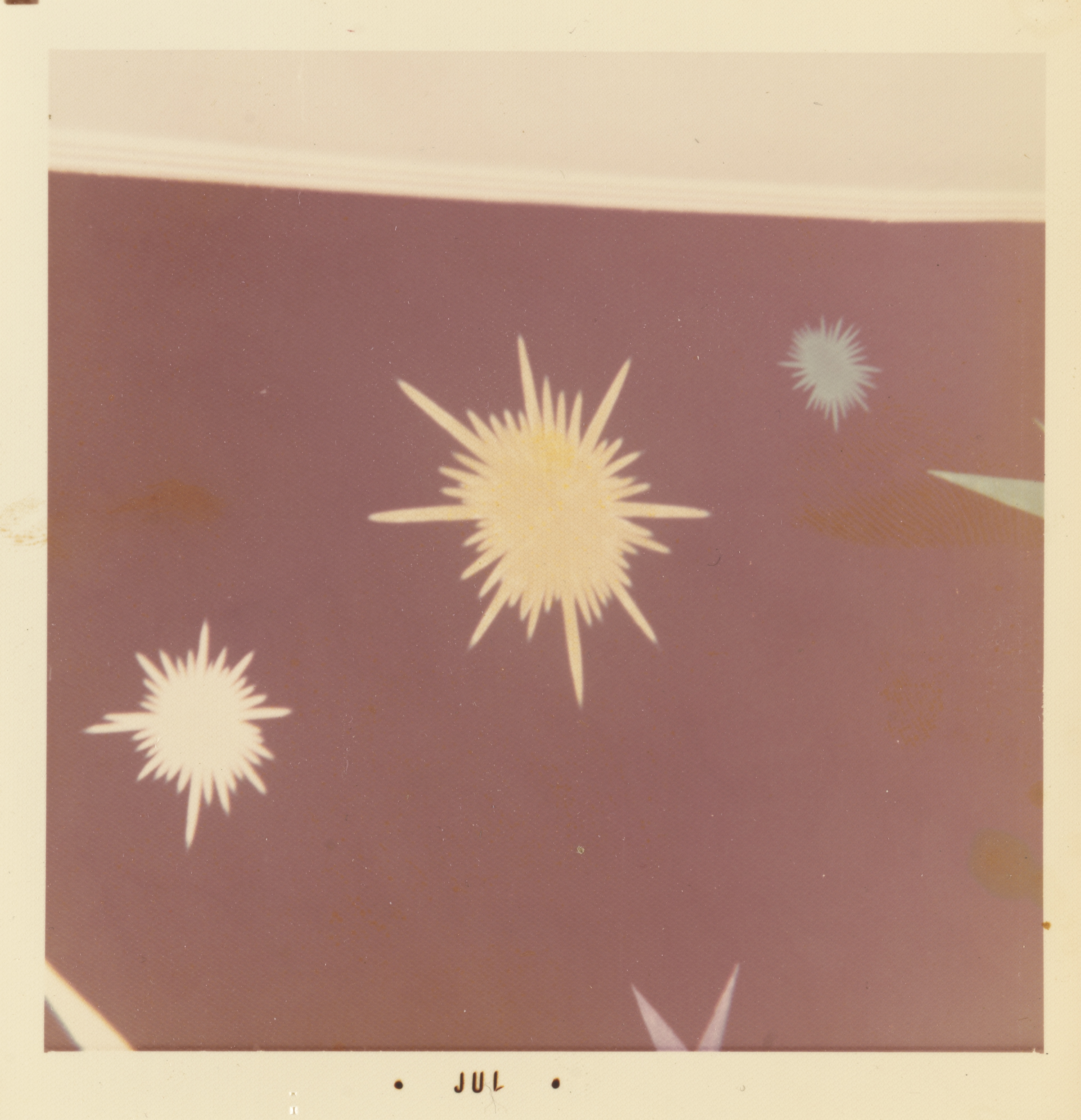 Photograph of a painted mural featuring stylized starburst forms on a mauve background. A large yellow burst with pointed rays is centered near the top, with a smaller white burst at lower left and a pale blue one at upper right. The edge of a ceiling molding is faintly visible along the upper edge. The white border of the print includes the text “• JUL •” centered along the bottom edge.