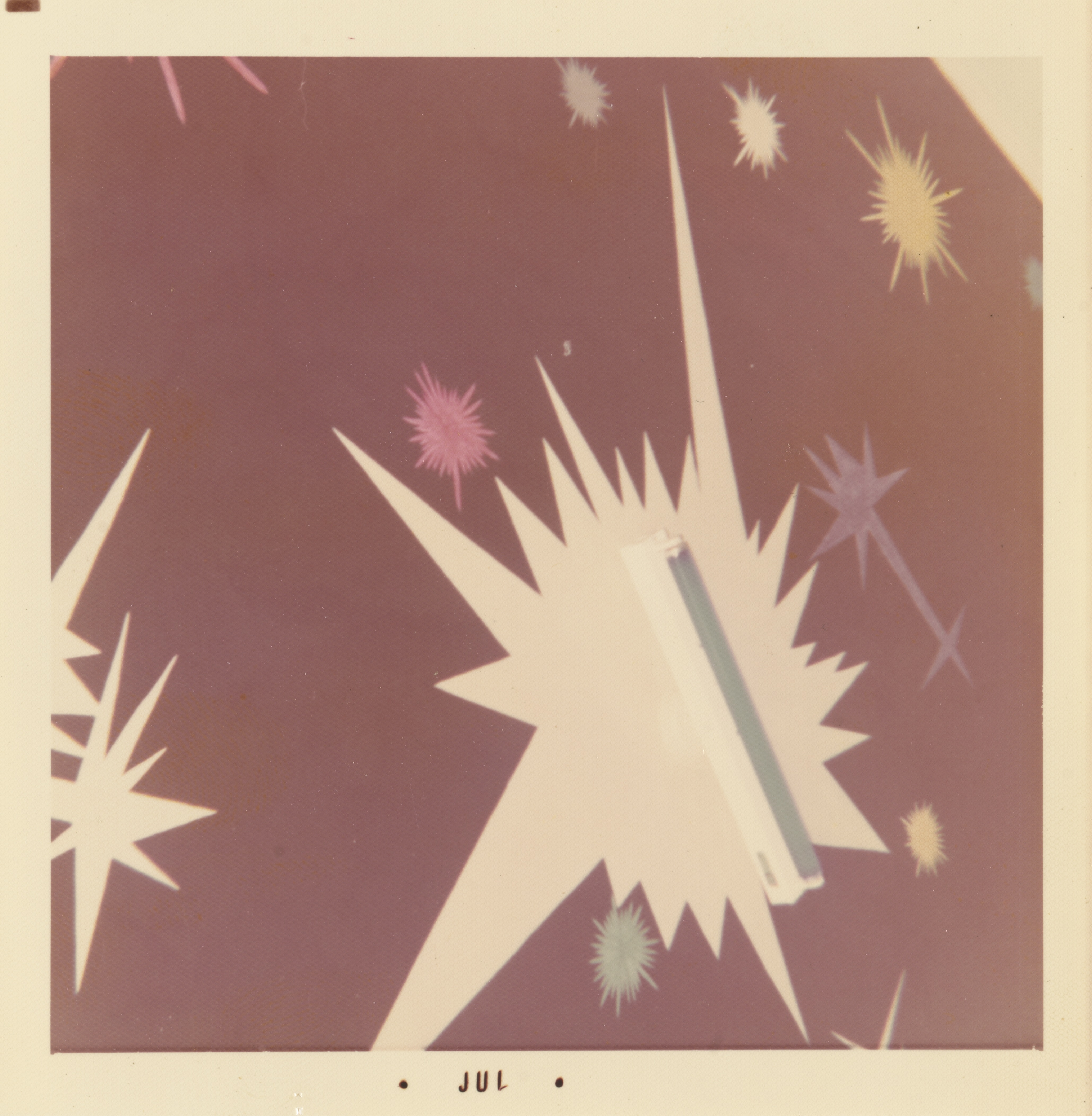 Photograph of a mural with a cosmic burst design on a mauve background. The composition features jagged starburst shapes in white, pink, yellow, green, and purple, radiating in various directions. At the center, a white fluorescent light fixture is mounted over a large white starburst, blending into the painted form. The white border of the print includes the text “• JUL •” centered along the bottom edge.